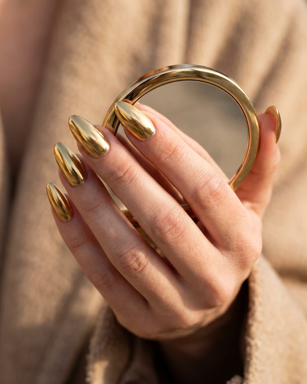 Long almond nails in highly reflective mirror gold chrome, holding a gold bangle against a warm beige background
