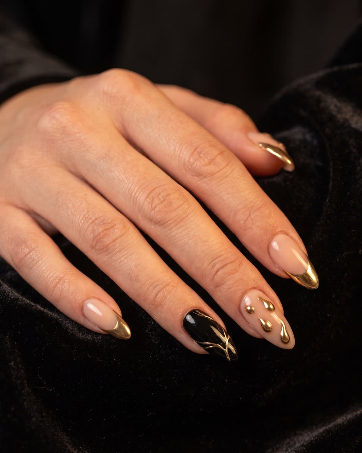Almond nails with sheer nude and gold chrome French tips, glossy black with gold chrome accents and nude with 3D gold chrome droplets, on black velvet