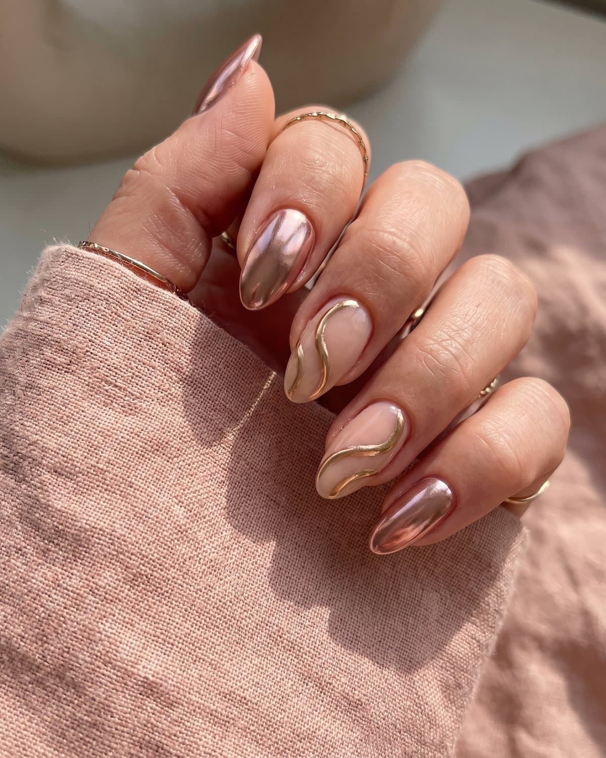 Almond nails with sheer nude and flowing gold chrome swirl lines and metallic rose gold chrome nails, on blush linen