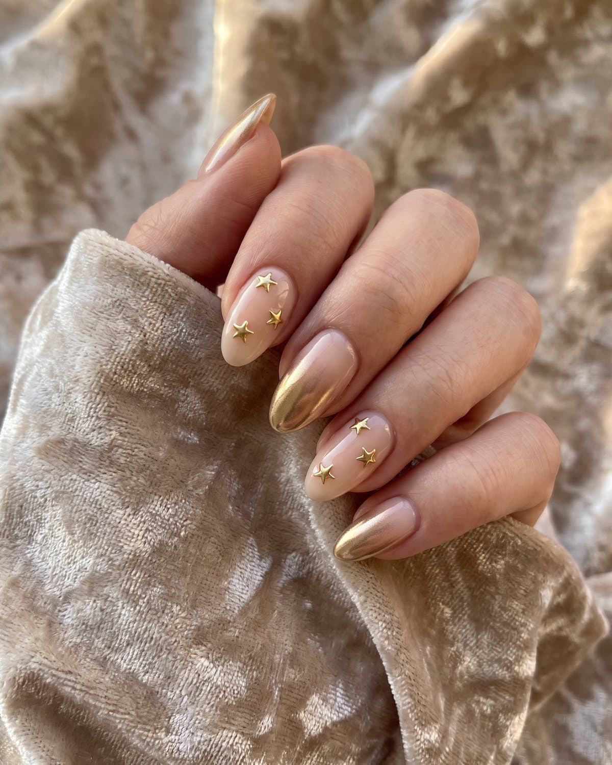 Almond nails with tiny raised gold chrome stars on nude and a gold chrome ombré from nude to mirror gold, on beige velvet
