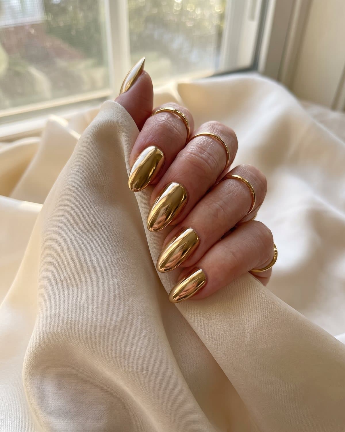 Almond nails in full mirror gold chrome like polished jewelry, with gold rings, resting on cream silk