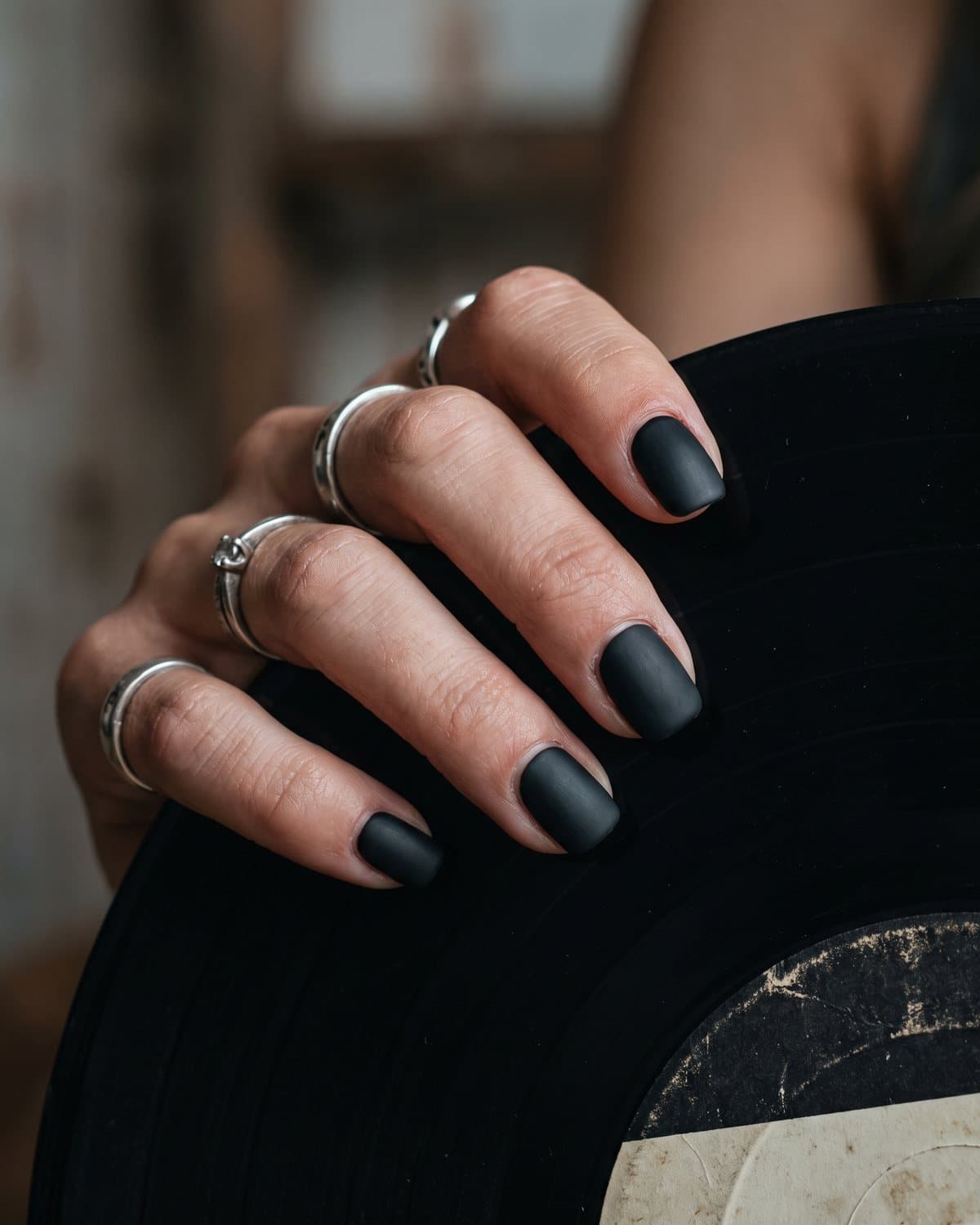 Short squoval nails in flat matte black, wearing silver rings and holding a vinyl record