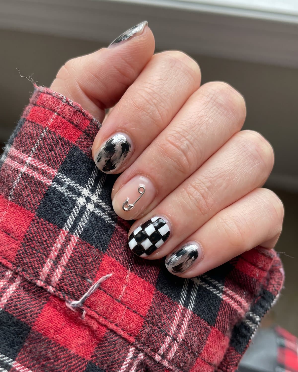 Short nails with smudged black and silver, a nude nail with a tiny silver safety pin charm and a black and white checkerboard nail, on a plaid flannel shirt