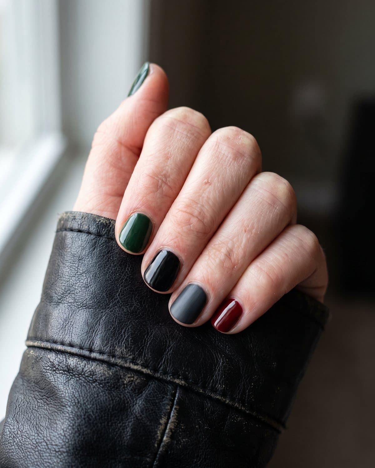 Short nails in glossy dark forest green mixed with black, charcoal grey and oxblood, on a black leather jacket
