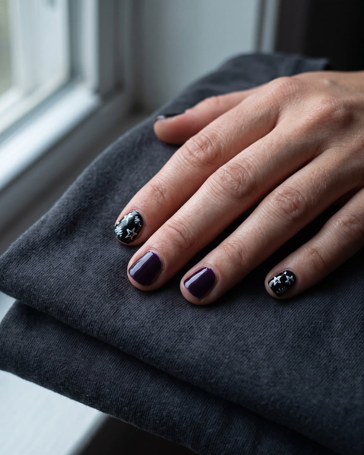 Short nails in deep dark purple and black nails with hand-drawn white stars and scribble doodles