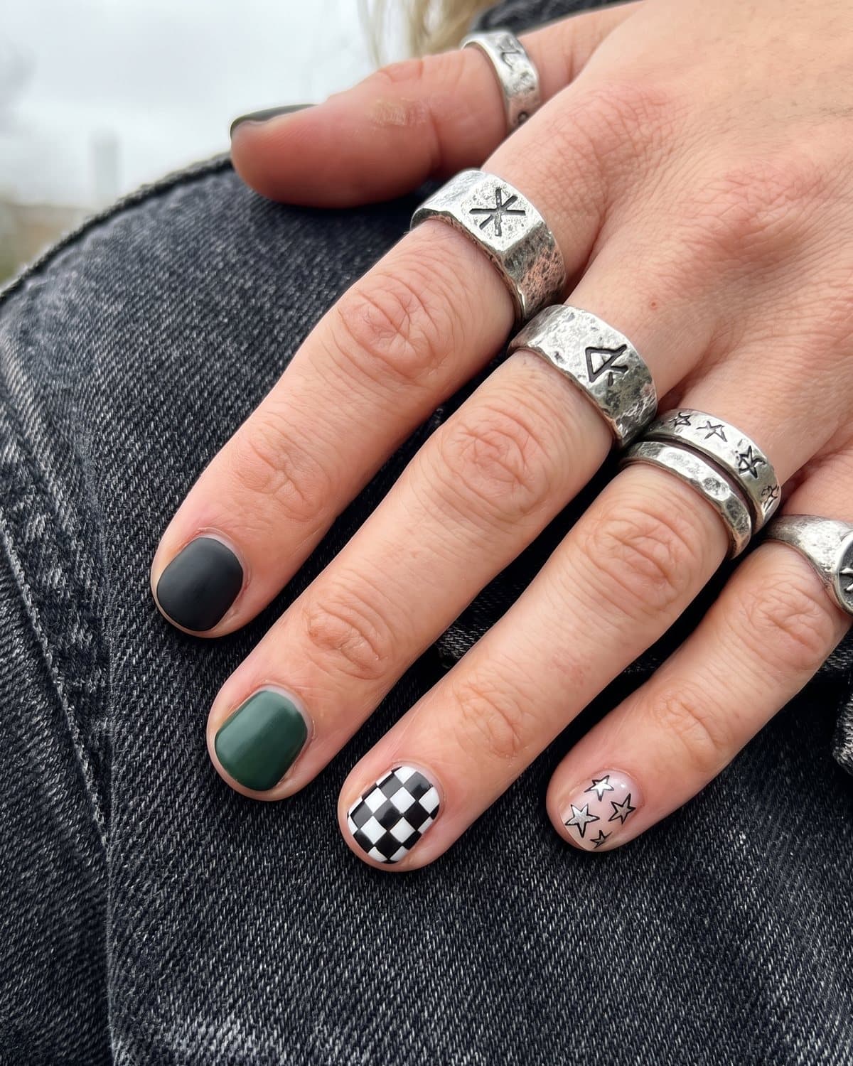 Short mismatched grunge nails in matte black, forest green, deep purple, checkerboard and silver star doodles, with chunky silver rings on black denim