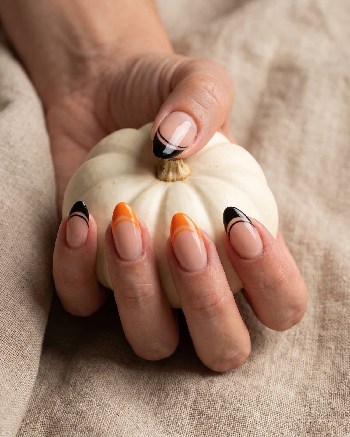 Almond nails with a sheer nude base and alternating black and orange French tips, holding a small white pumpkin