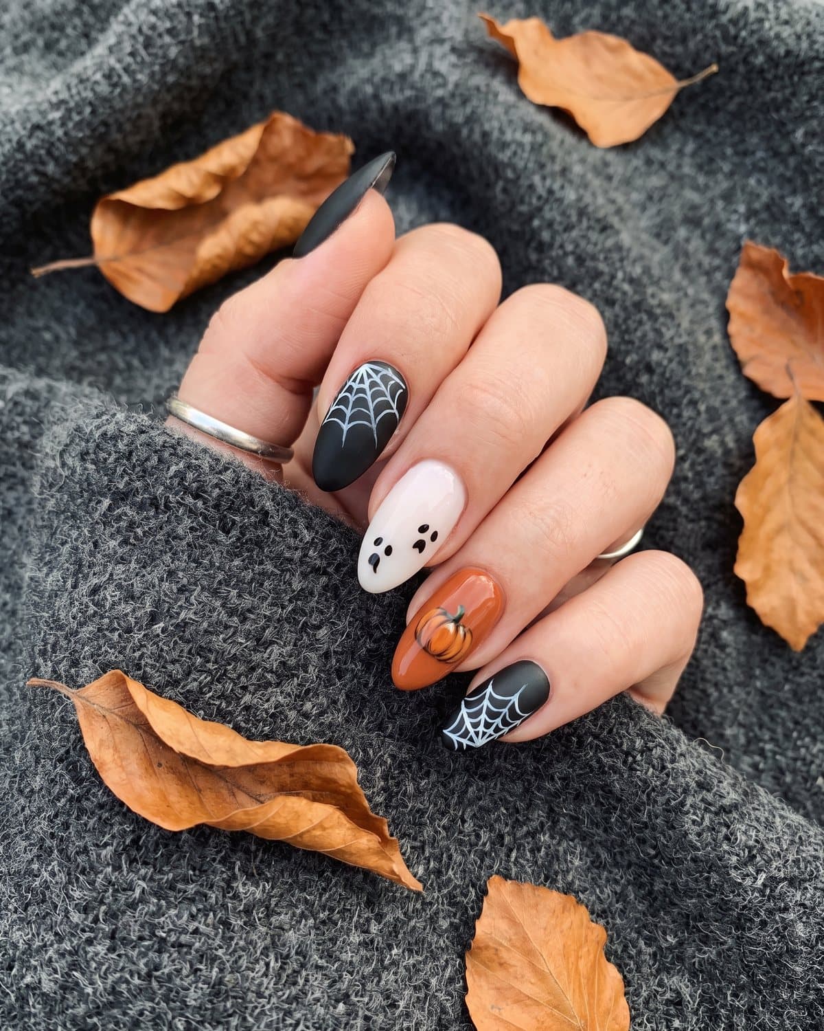 Matte black almond nails with white spider webs, milky white nails with tiny ghost faces and an orange pumpkin nail