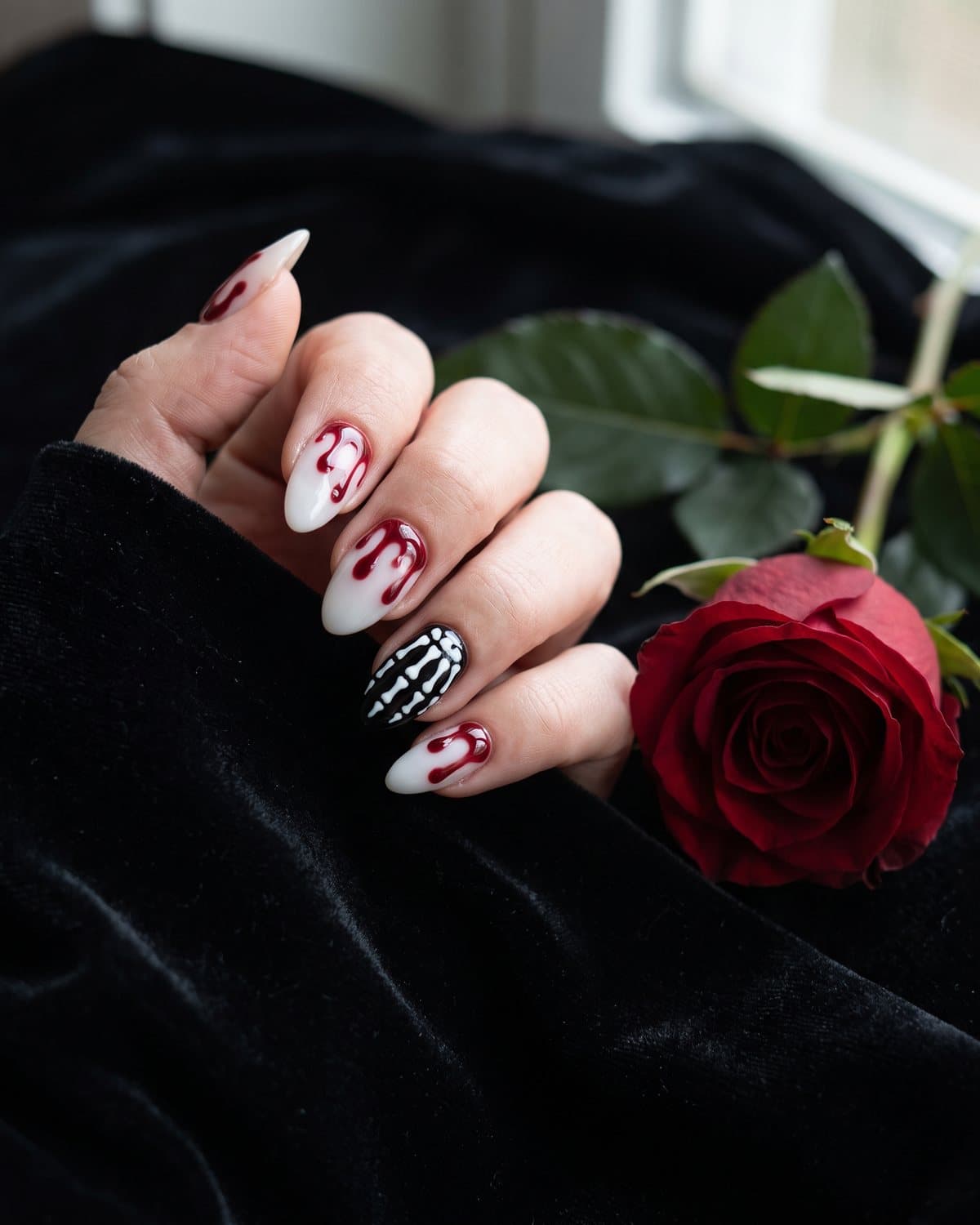 Milky white almond nails with glossy red blood drips and black nails with white skeleton bone art, resting on black velvet with a red rose