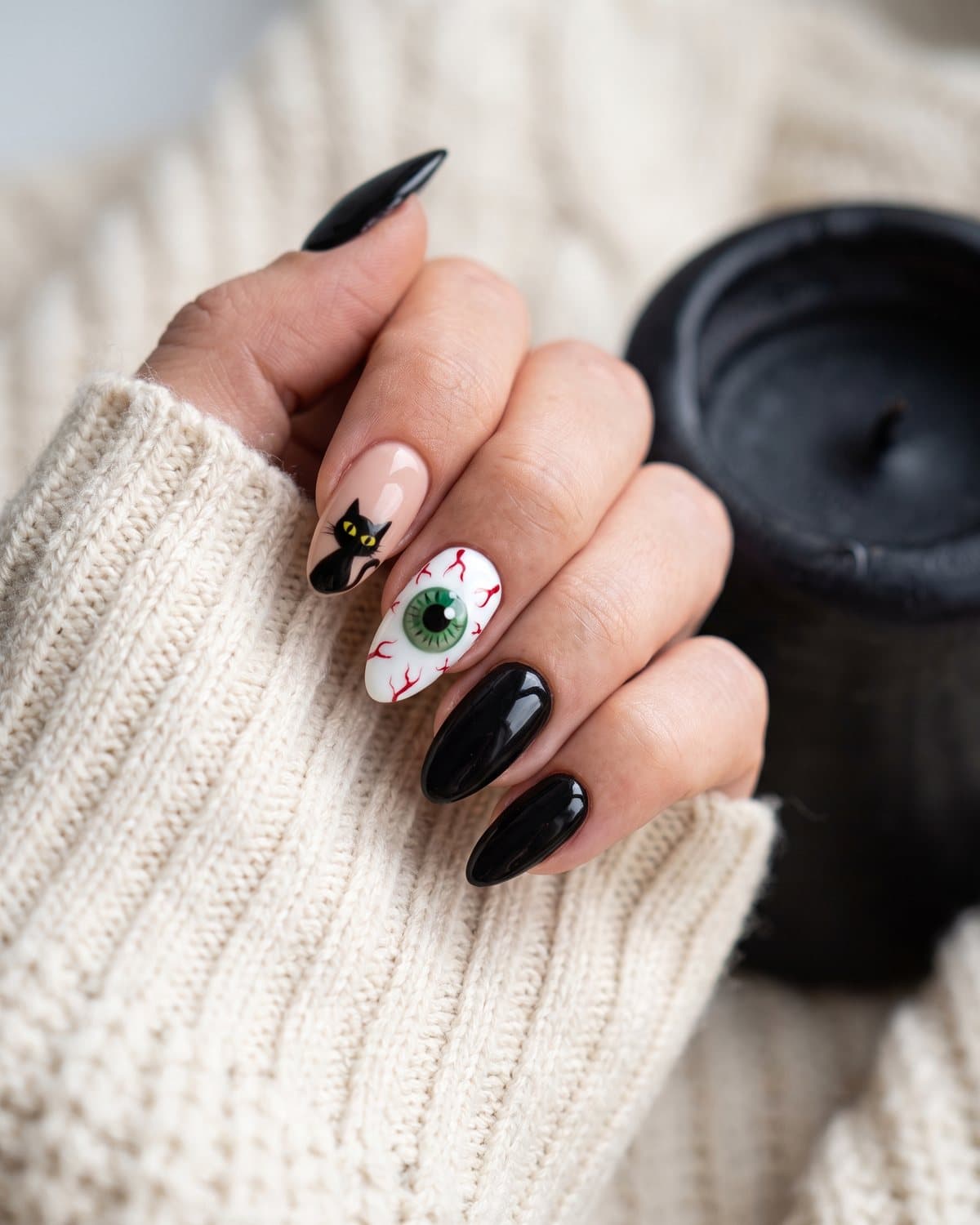 Glossy almond nails with a black cat on a nude nail, a realistic green eyeball nail and solid black nails, on a cream knit sweater