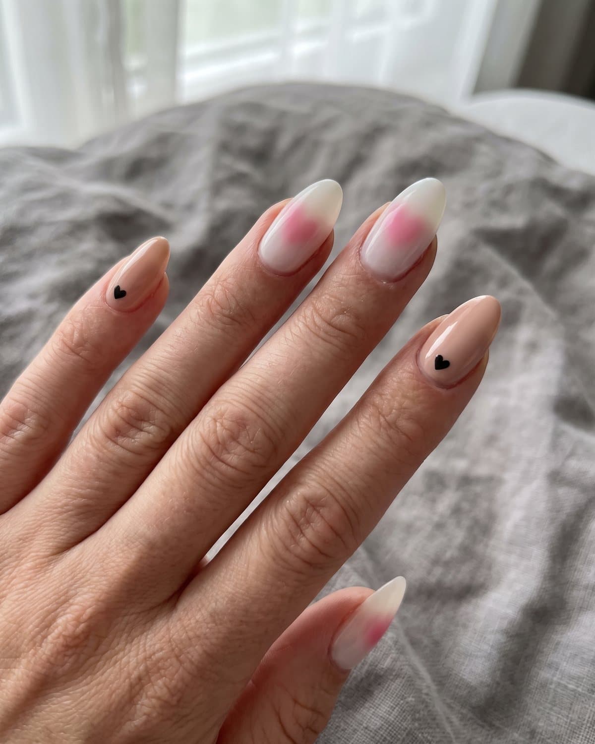 Almond nails with small glossy black hearts on nude and milky nails with a soft blurred pink heart aura, on grey linen