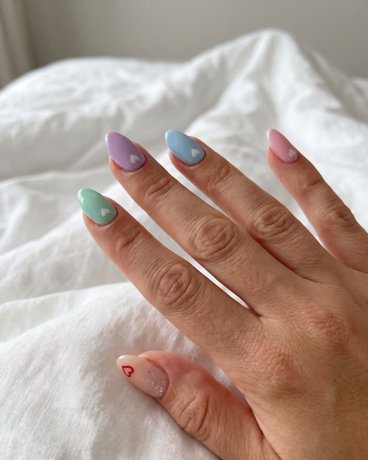 Almond nails with mismatched pastel candy hearts in mint, lilac, baby blue and pink with small white hearts, and nude nails with a red heart and fine glitter