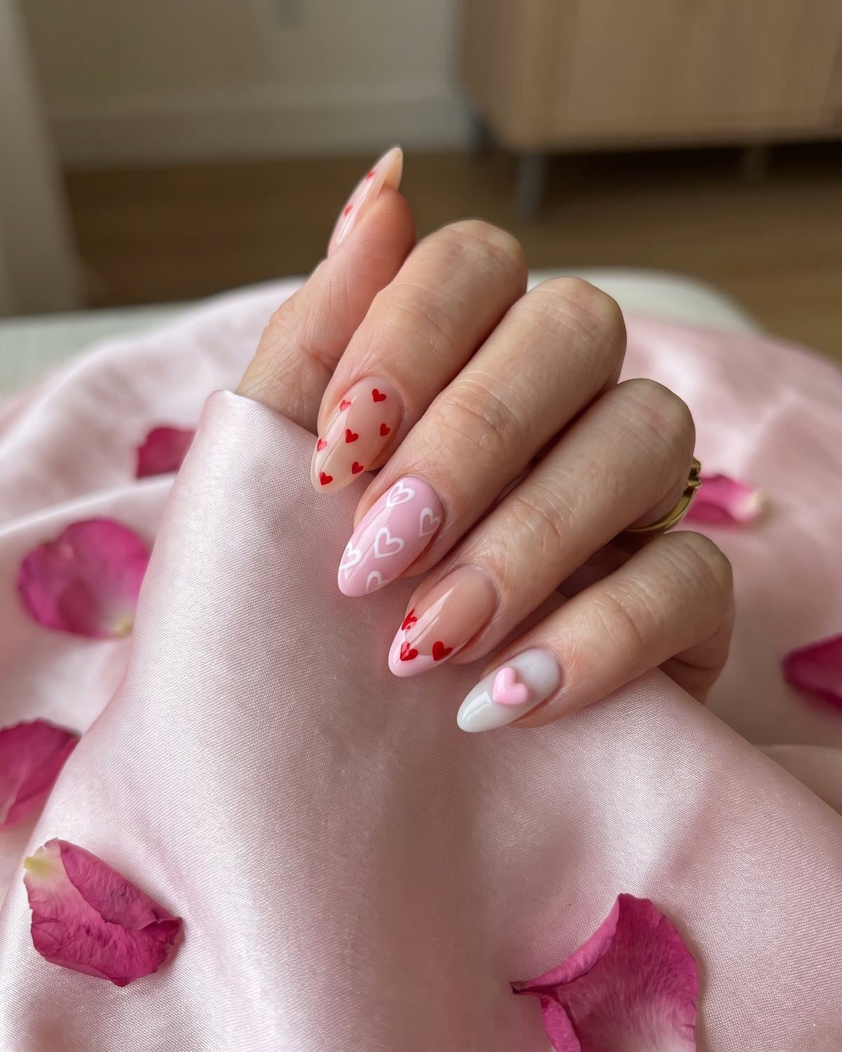 Almond heart nails with tiny red hearts, white heart outlines, red heart French tips and a 3D pink heart charm, on pale pink satin with rose petals