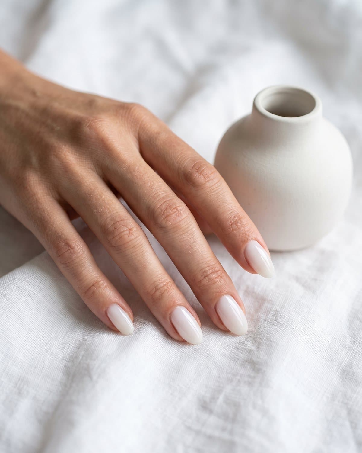 Glossy oval nails in a classic milky white with a soft translucent finish, resting on white linen beside a white ceramic vase