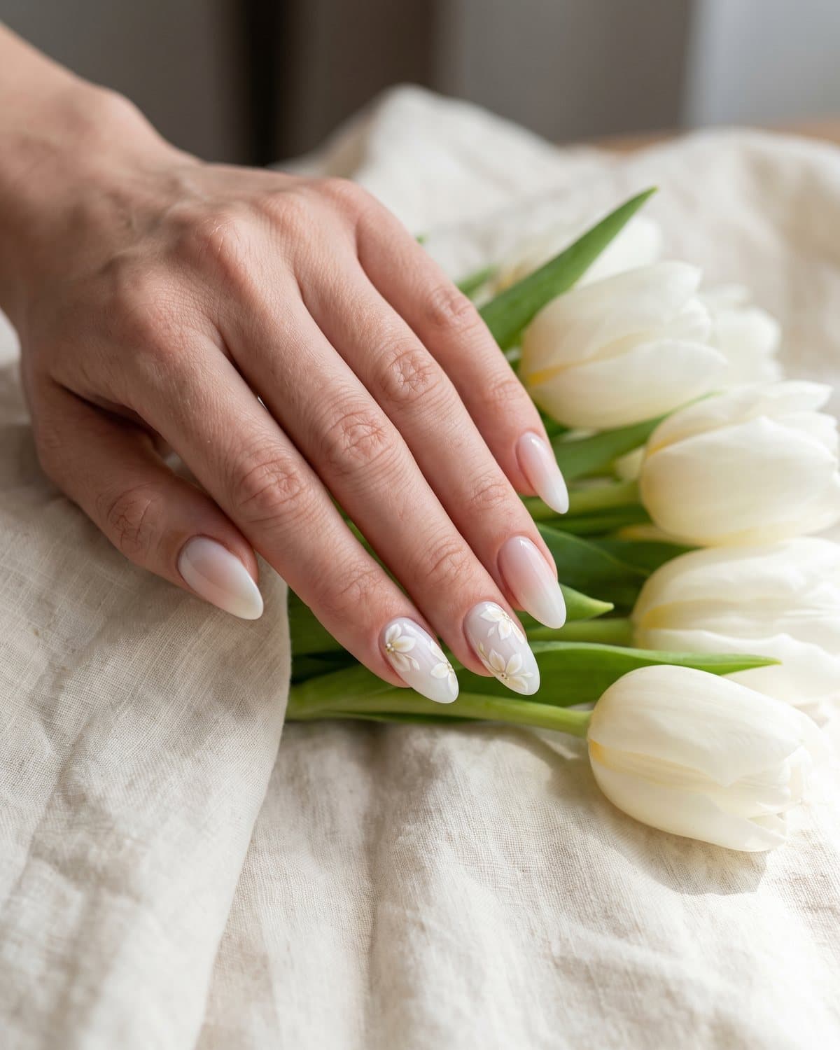 Almond nails with milky white and tiny white and pale yellow flowers, and a milky white to nude ombré, beside white tulips