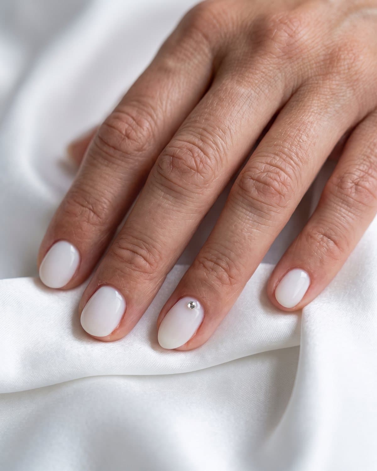 Short round milky white nails with one milky white almond accent with a small rhinestone near the cuticle, on white satin