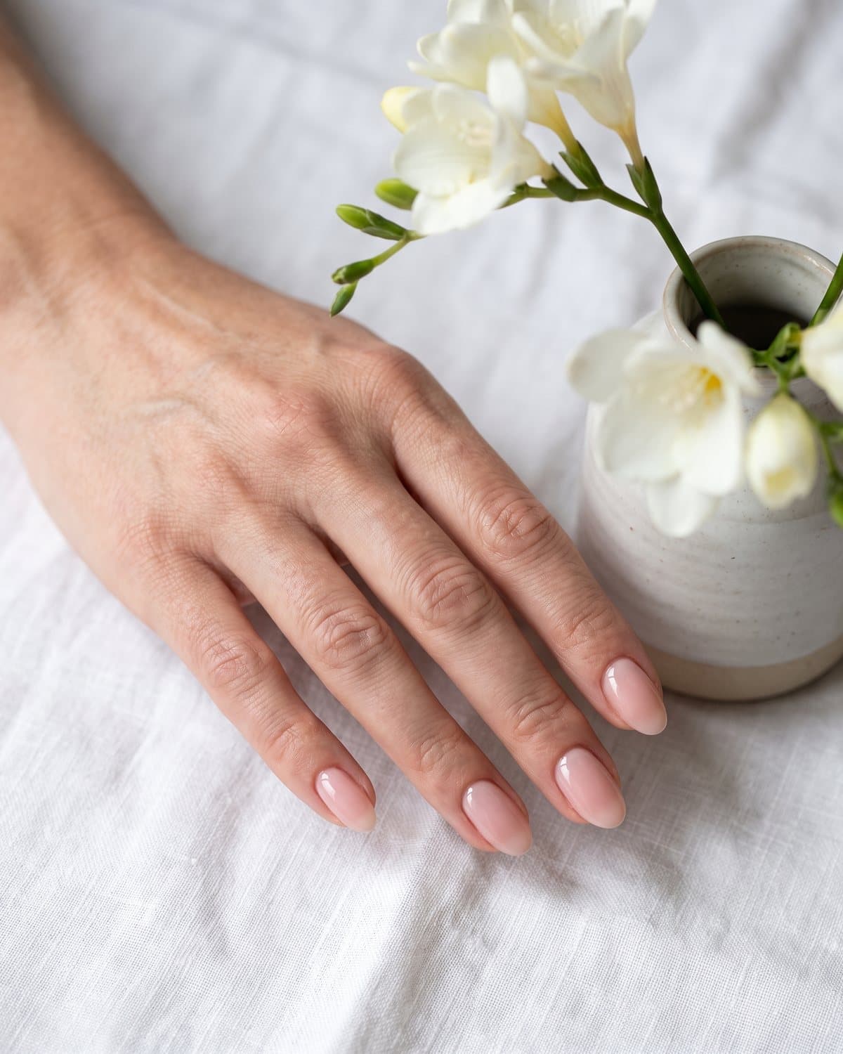Fair-skinned hand with glossy oval nails in a sheer pink nude, resting on white linen next to white flowers