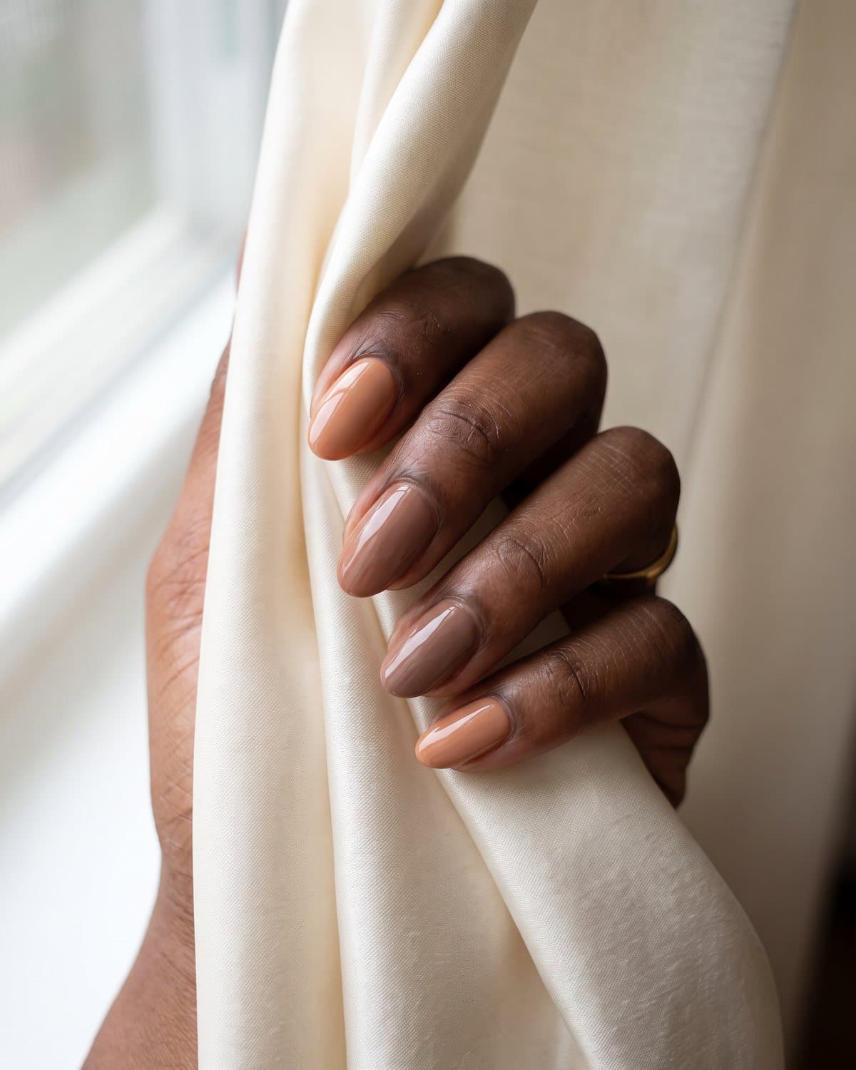 Deep brown skin with glossy oval nails alternating between rich caramel nude and warm mocha nude, on cream silk with a gold ring