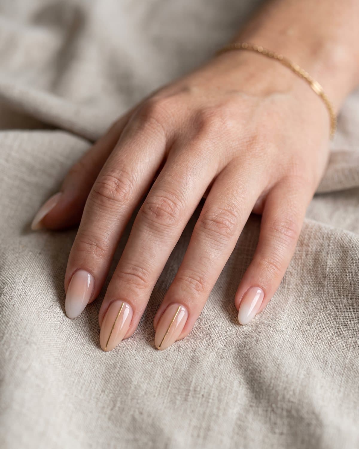 Almond nude nails with fine gold vertical lines and a soft nude-to-milky-white ombré, on beige linen with a gold bracelet