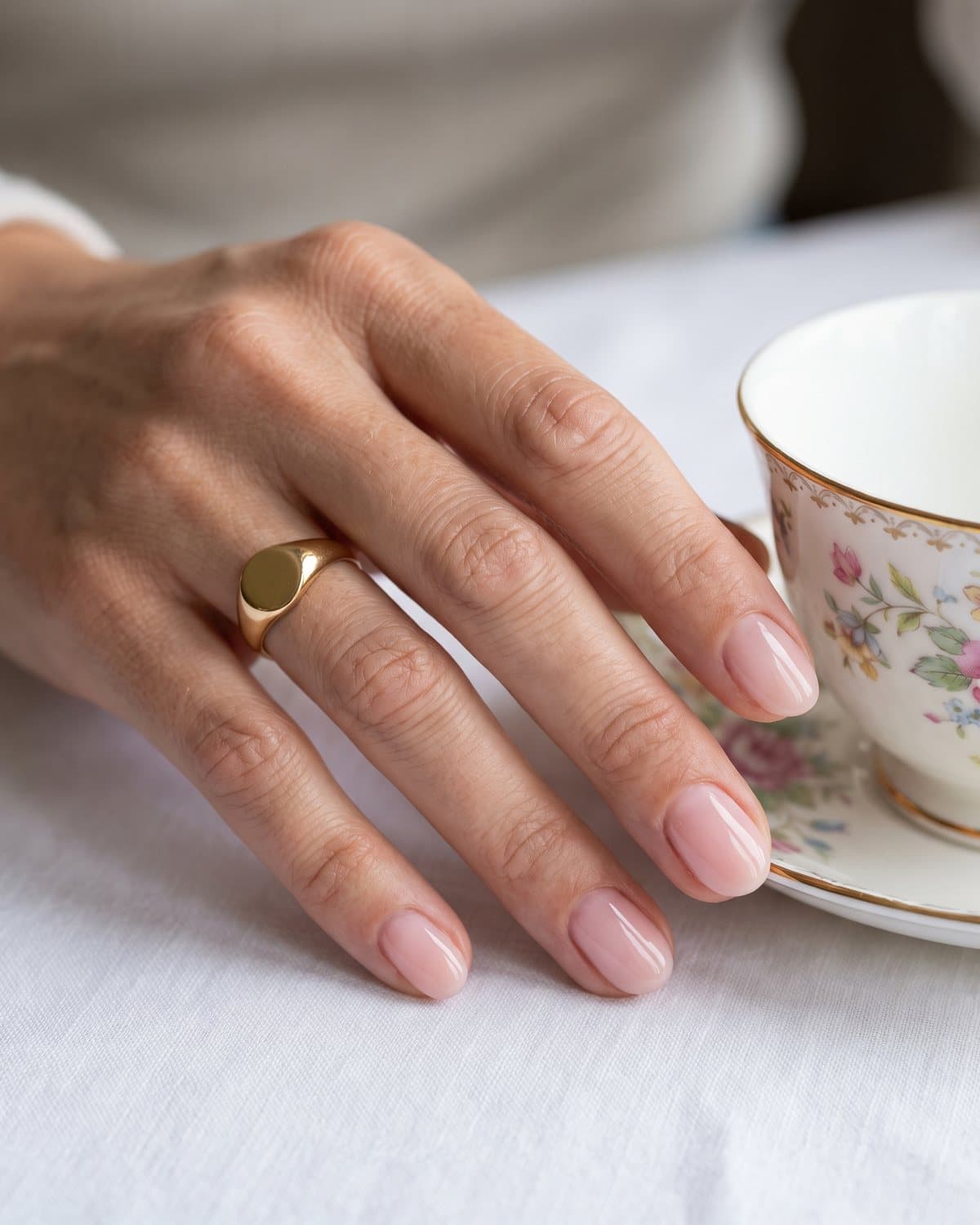Short oval nails in a sheer glossy ballet pink, with a gold signet ring, beside a porcelain teacup on a white tablecloth