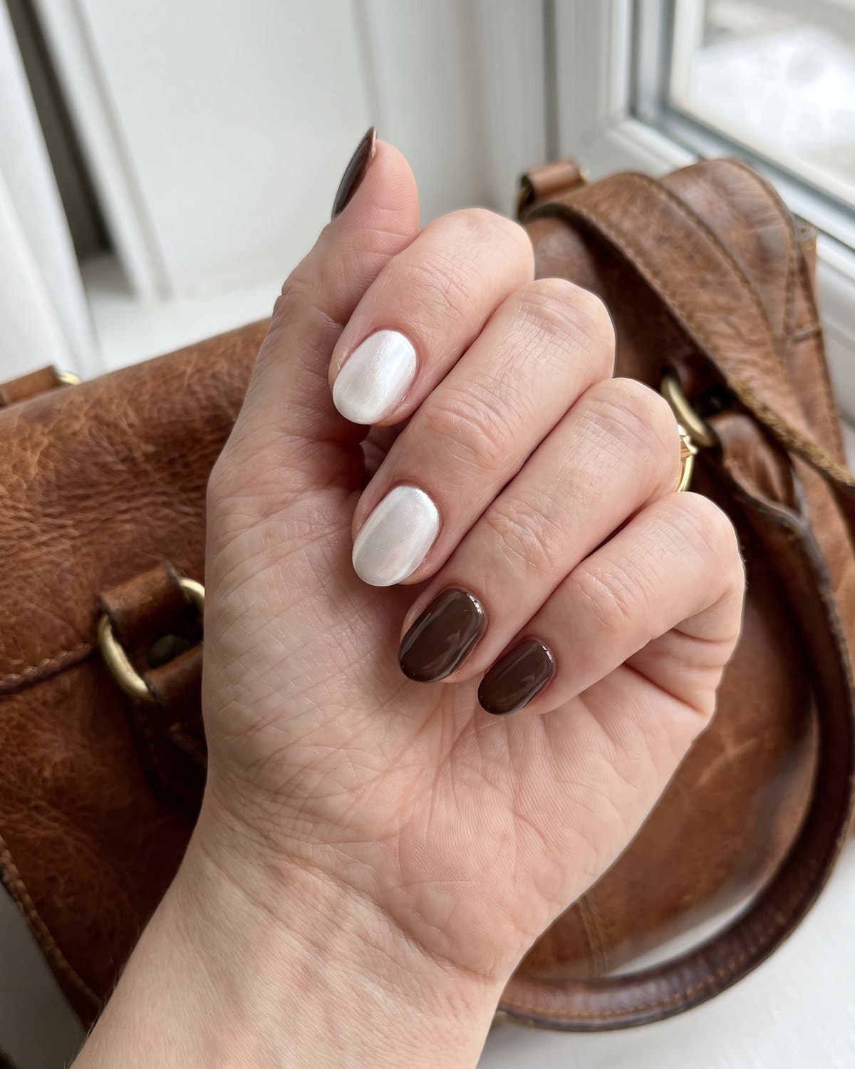 Short oval nails in soft pearl chrome white and glossy espresso brown, resting on a brown leather handbag