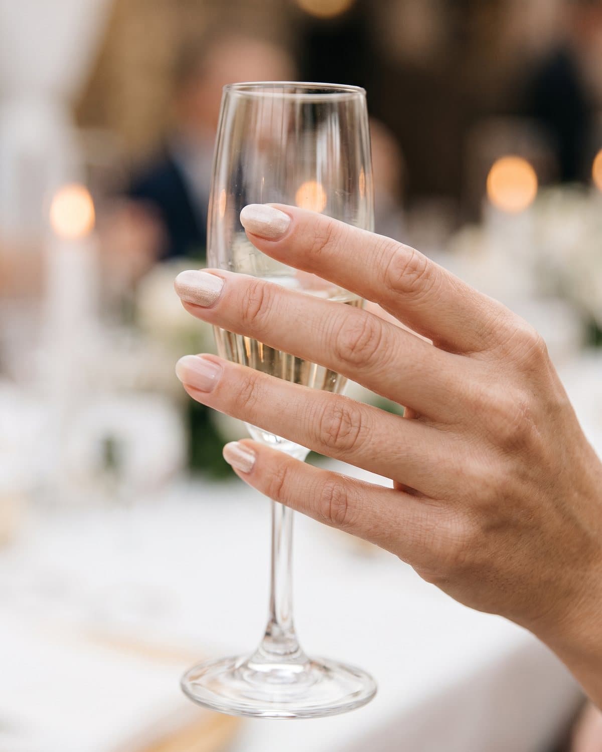 Short oval nails in soft champagne shimmer and natural buffed shiny nails, holding a champagne flute