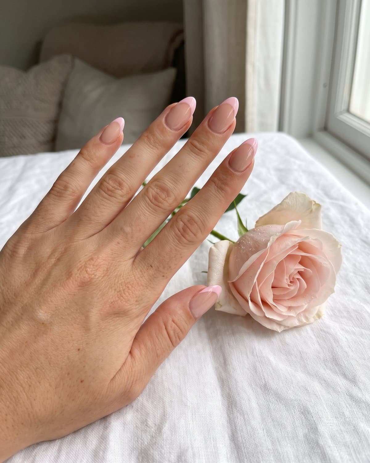 Glossy almond nails with a sheer nude base and soft baby pink French tips, resting on white linen with a pink rose