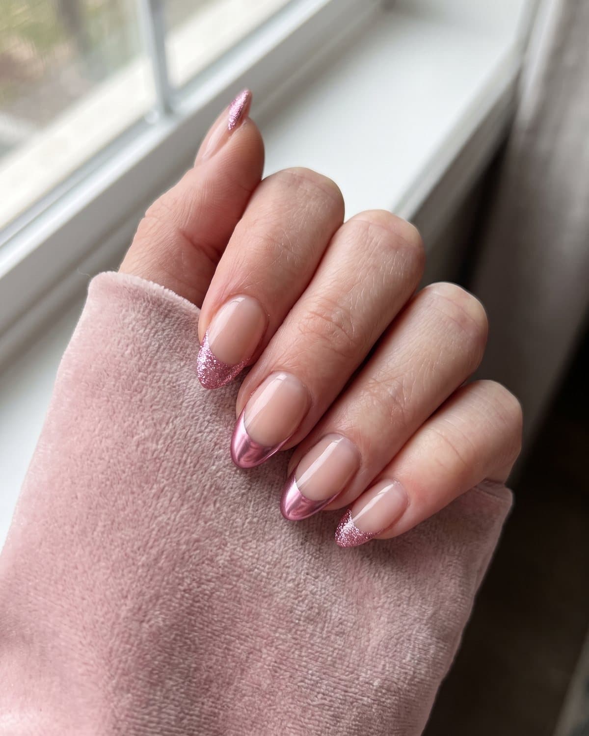 Almond nails with sheer nude bases and sparkly pink glitter French tips and metallic pink chrome French tips, on pale pink velvet