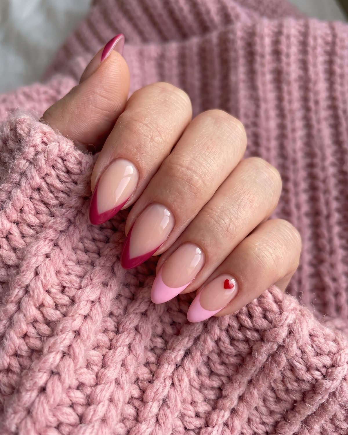 Almond nails with sheer nude bases and pointed pink V-shaped French tips, and pink French tips with a tiny red heart, on a pink knit