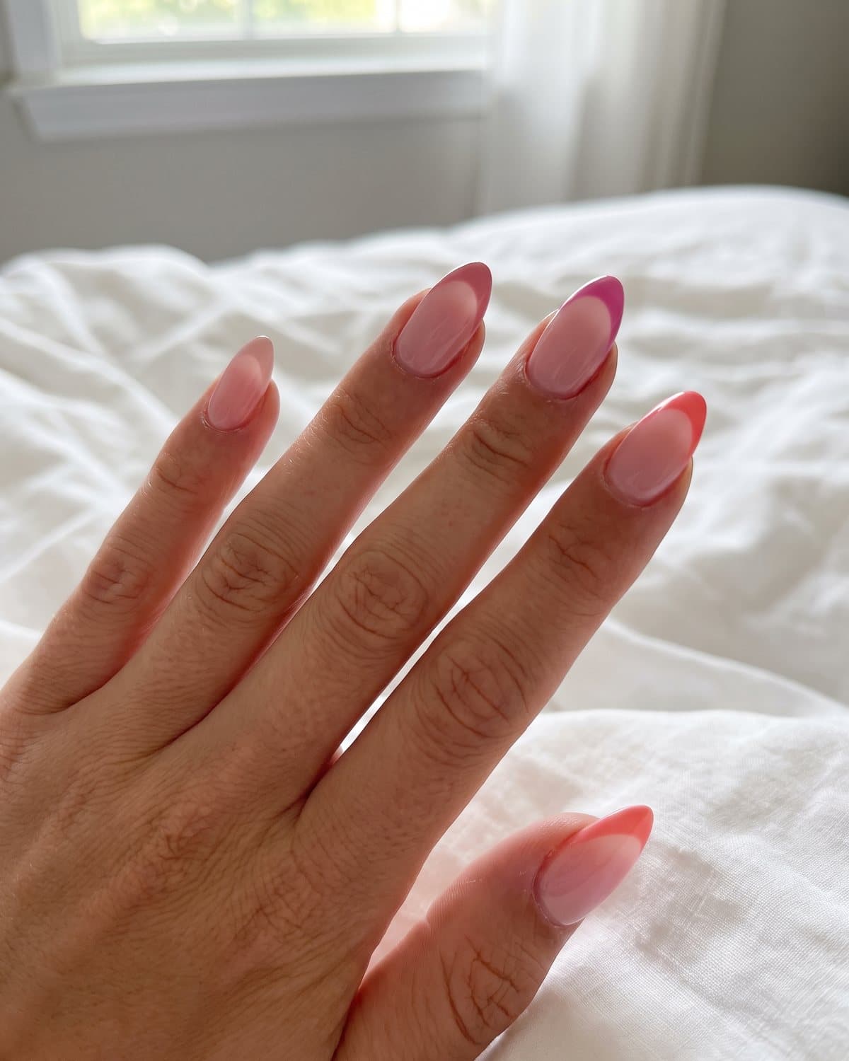 Almond nails with a soft pink ombré fading into the tip and mismatched pink French tips in blush, rose, fuchsia and coral, on white bedding