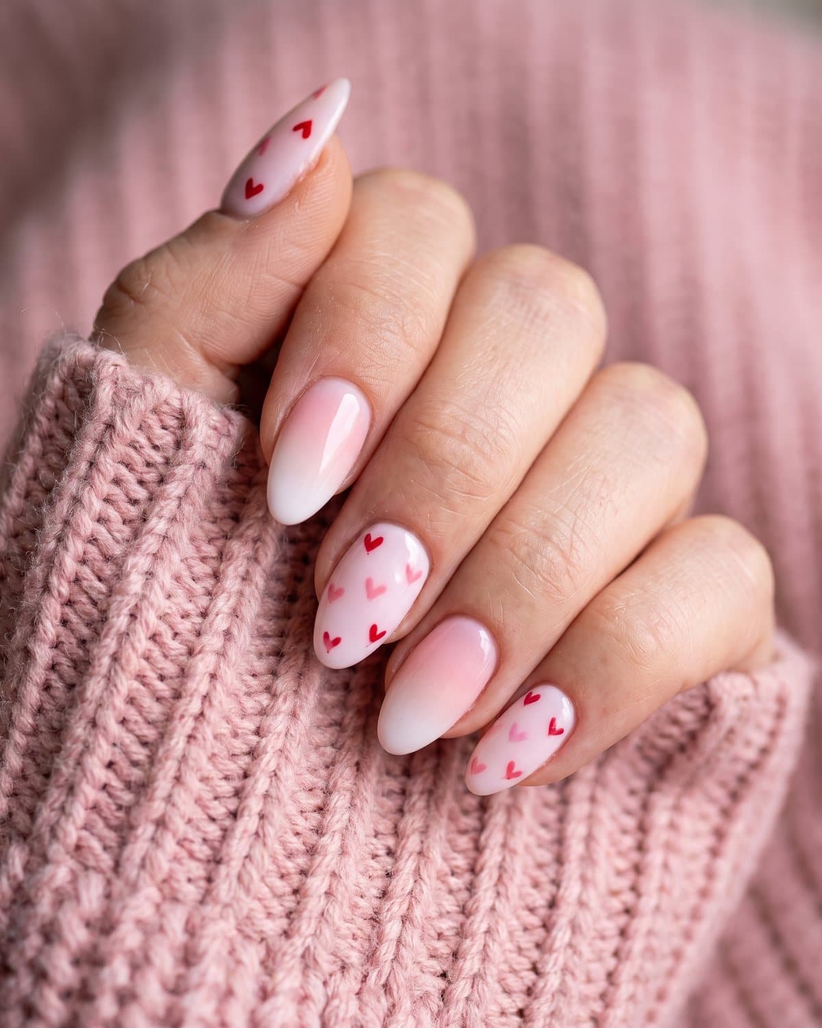 Almond nails with a soft pink-to-white baby boomer ombré and pale pink nails with tiny red and pink hearts, on a pink knit sweater