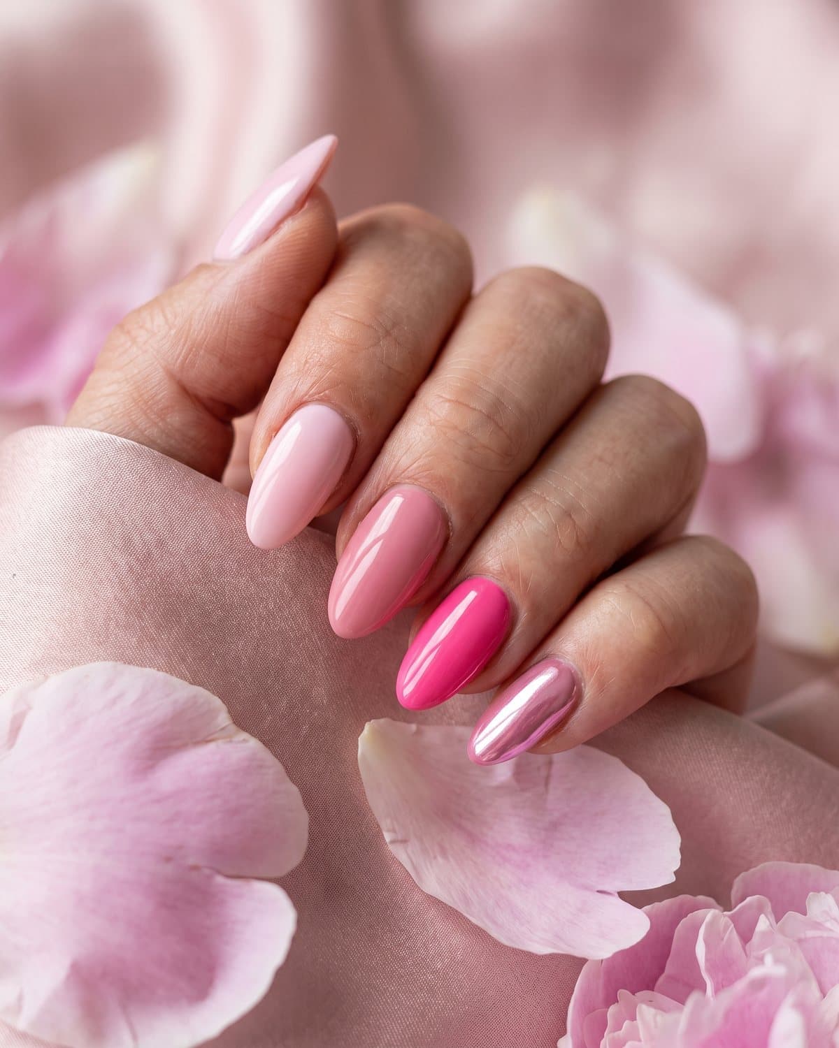 Glossy almond nails in a gradient of pink shades from baby pink to hot pink and pink chrome, on blush satin with peony petals