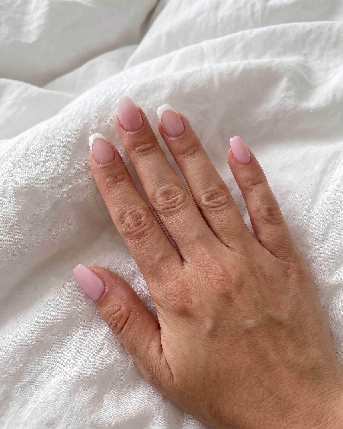 Short acrylic nails in soft pink coffin, short almond with white French tips and pink-and-white ombré, on white bedding