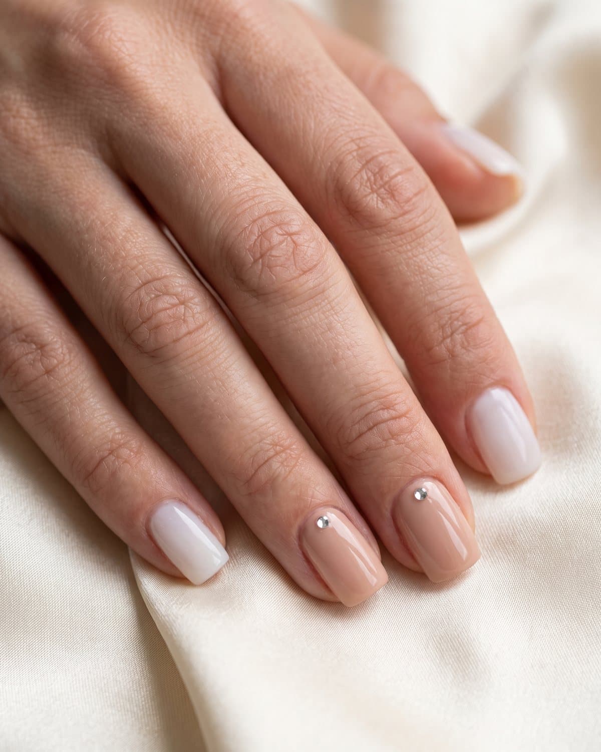 Short acrylic nails in milky white and nude with a single small rhinestone near the cuticle, on cream satin