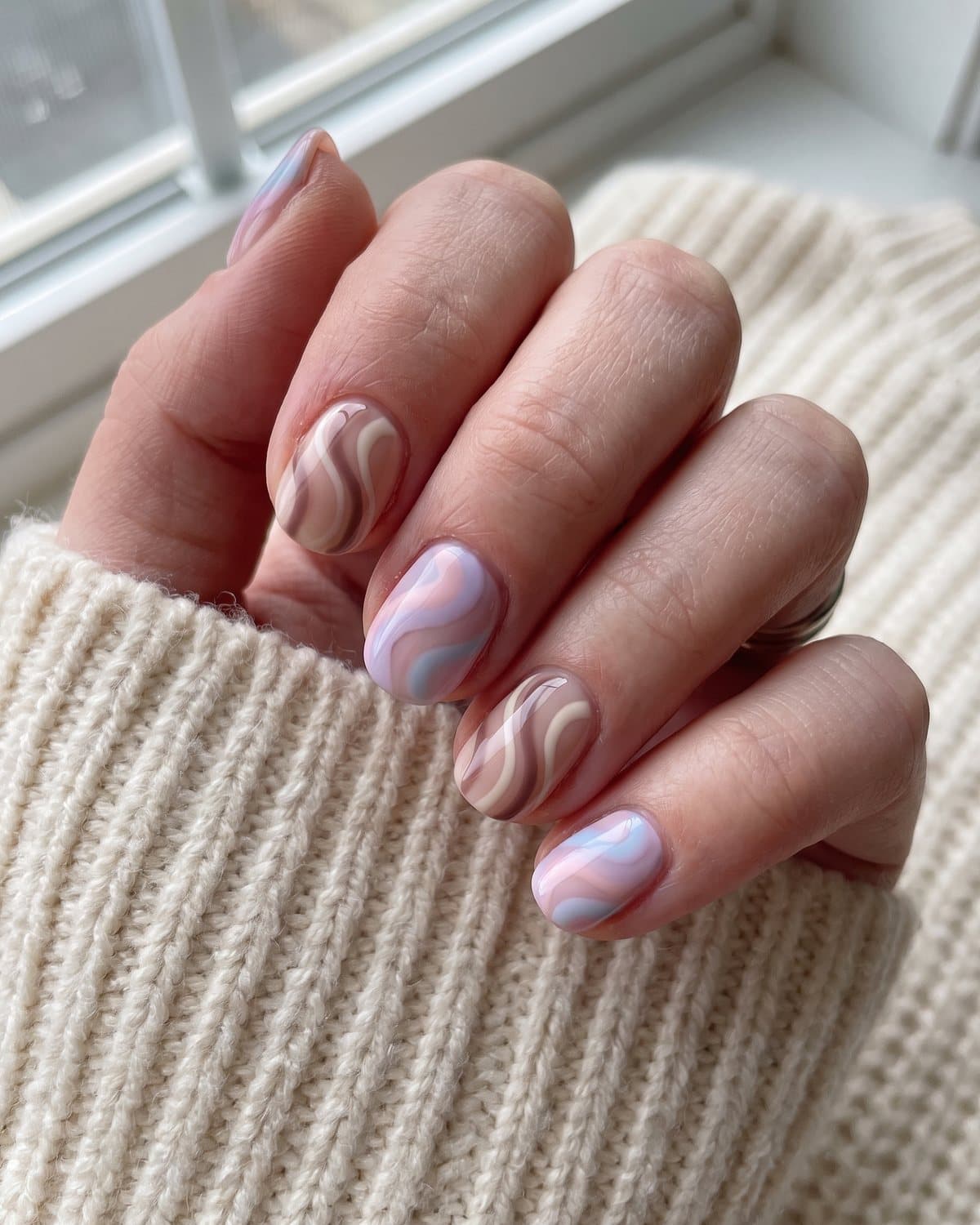 Short acrylic nails with abstract brown and cream lines and soft pastel swirls in pink, lilac and blue on nude, on a cream knit