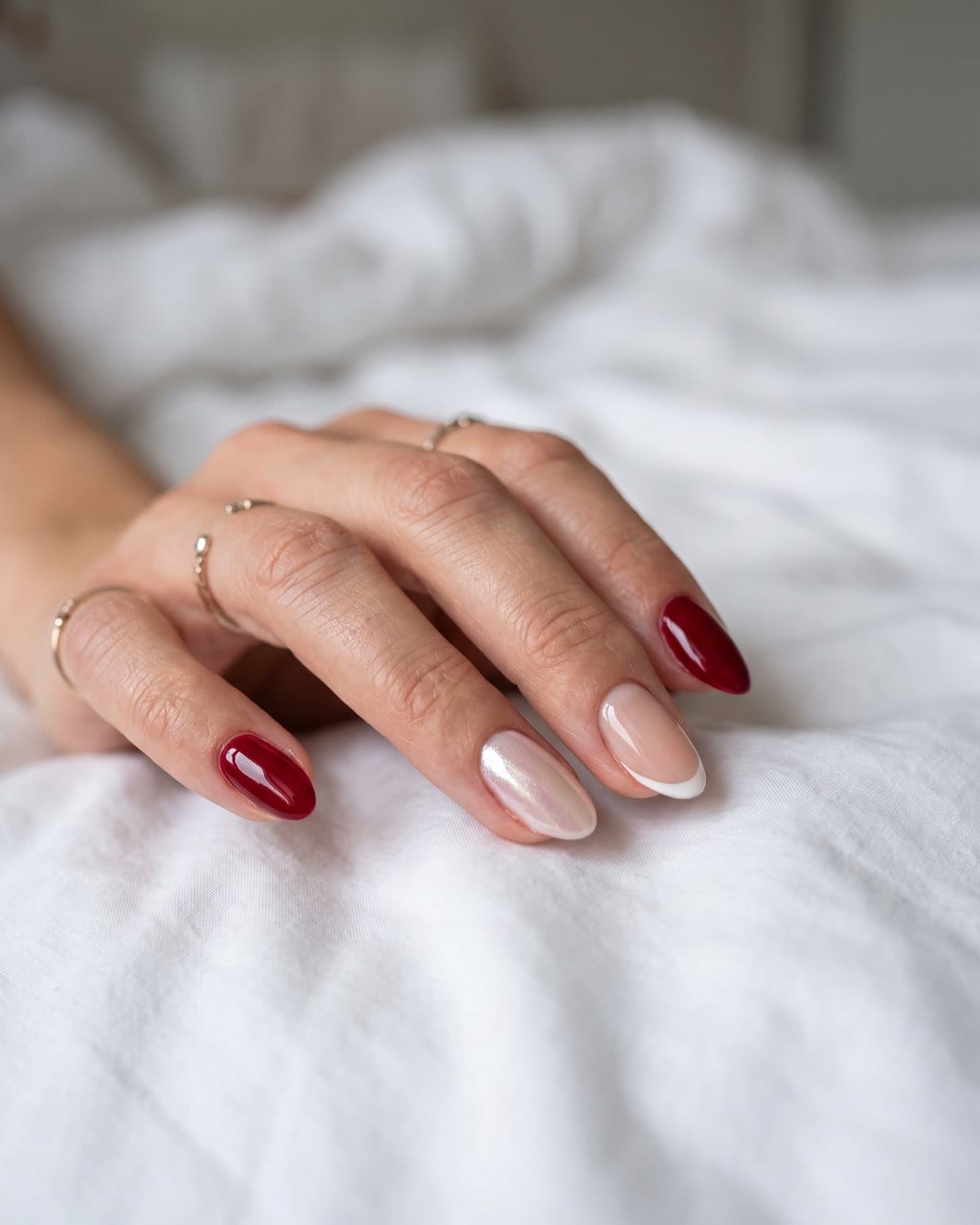 Short almond nails in deep cherry red alternating with sheer nude micro French tips and a pearly glazed nail, resting on white sheets