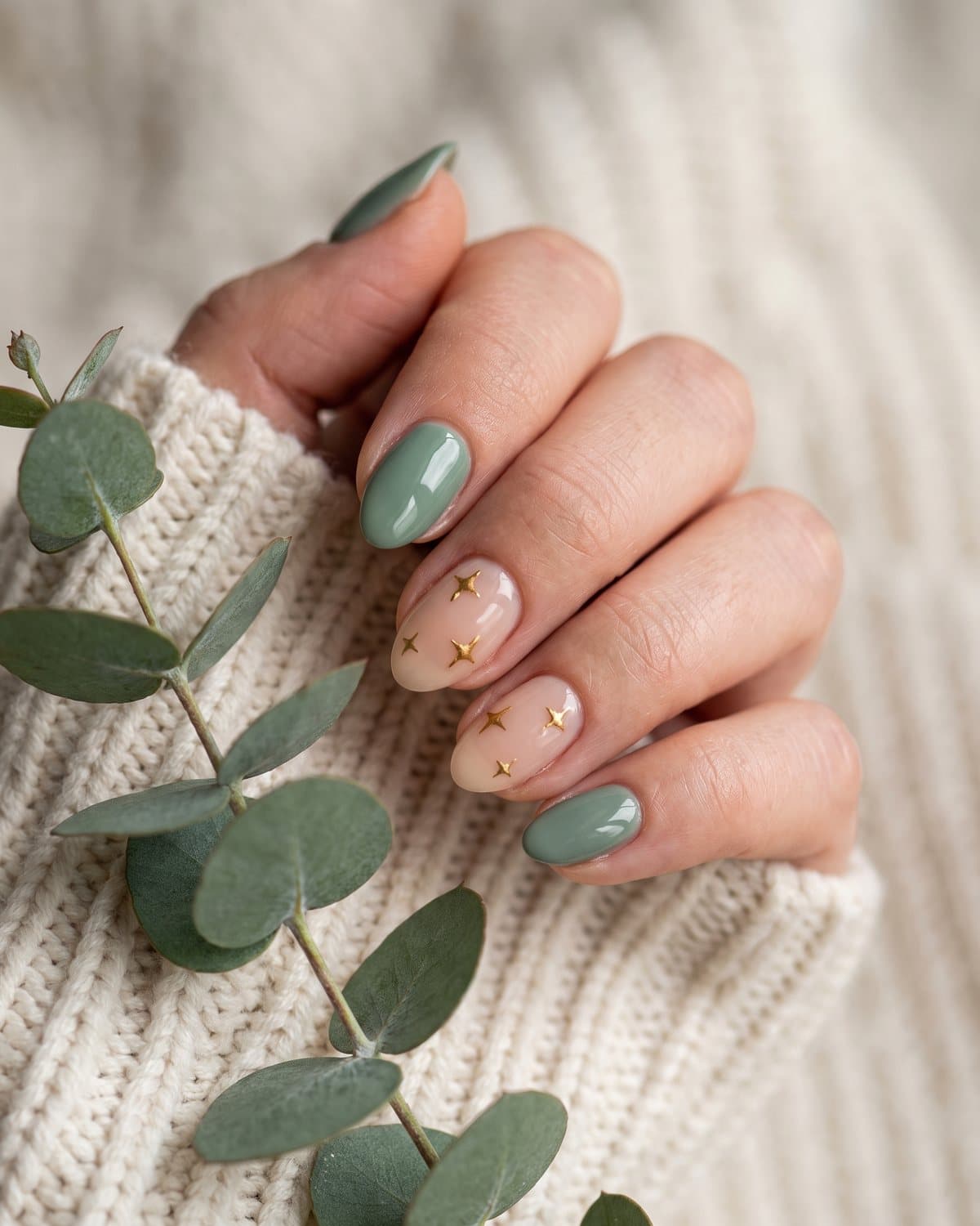 Short almond nails in glossy sage green and sheer nude with tiny gold stars, on a cream knit sweater with eucalyptus