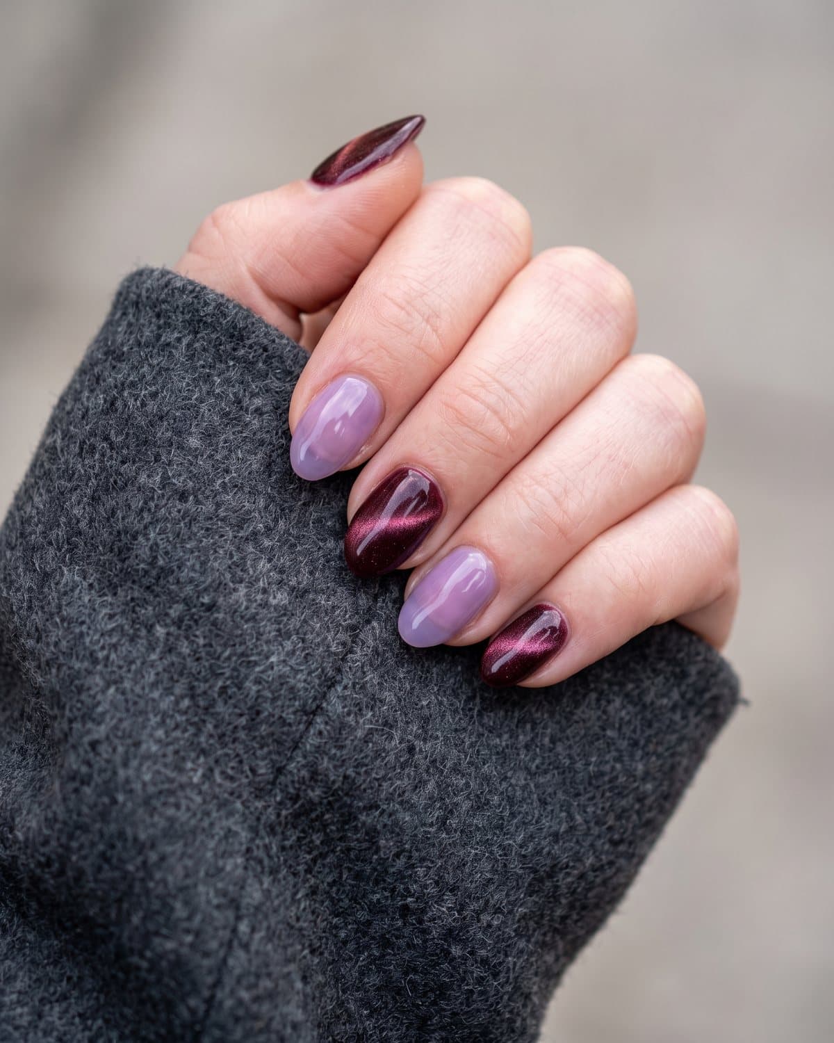 Short almond nails in burgundy cat-eye shimmer alternating with translucent lavender jelly, resting on a grey wool sleeve