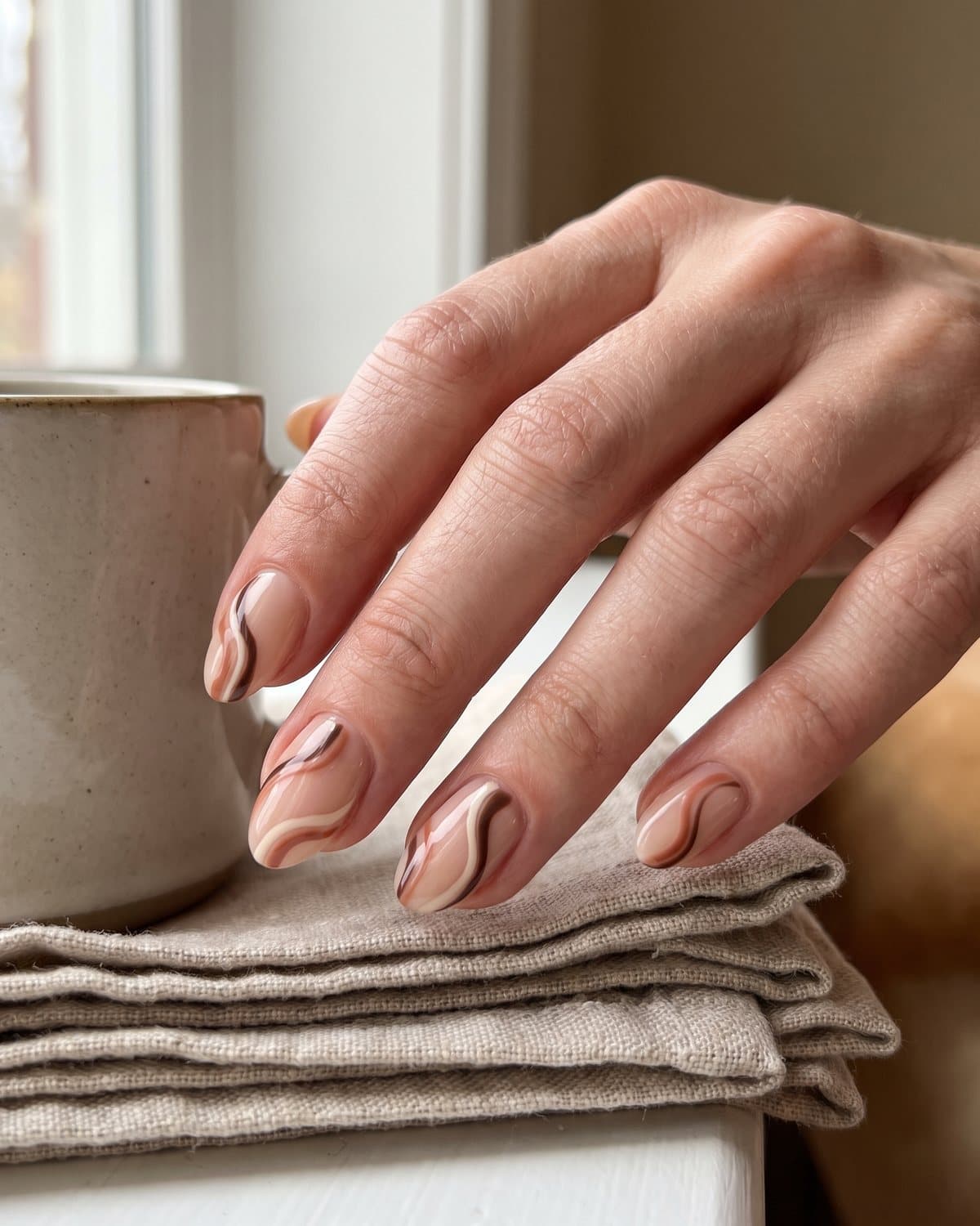 Short almond nails with a sheer nude base and abstract swirls in chocolate brown, cream and terracotta