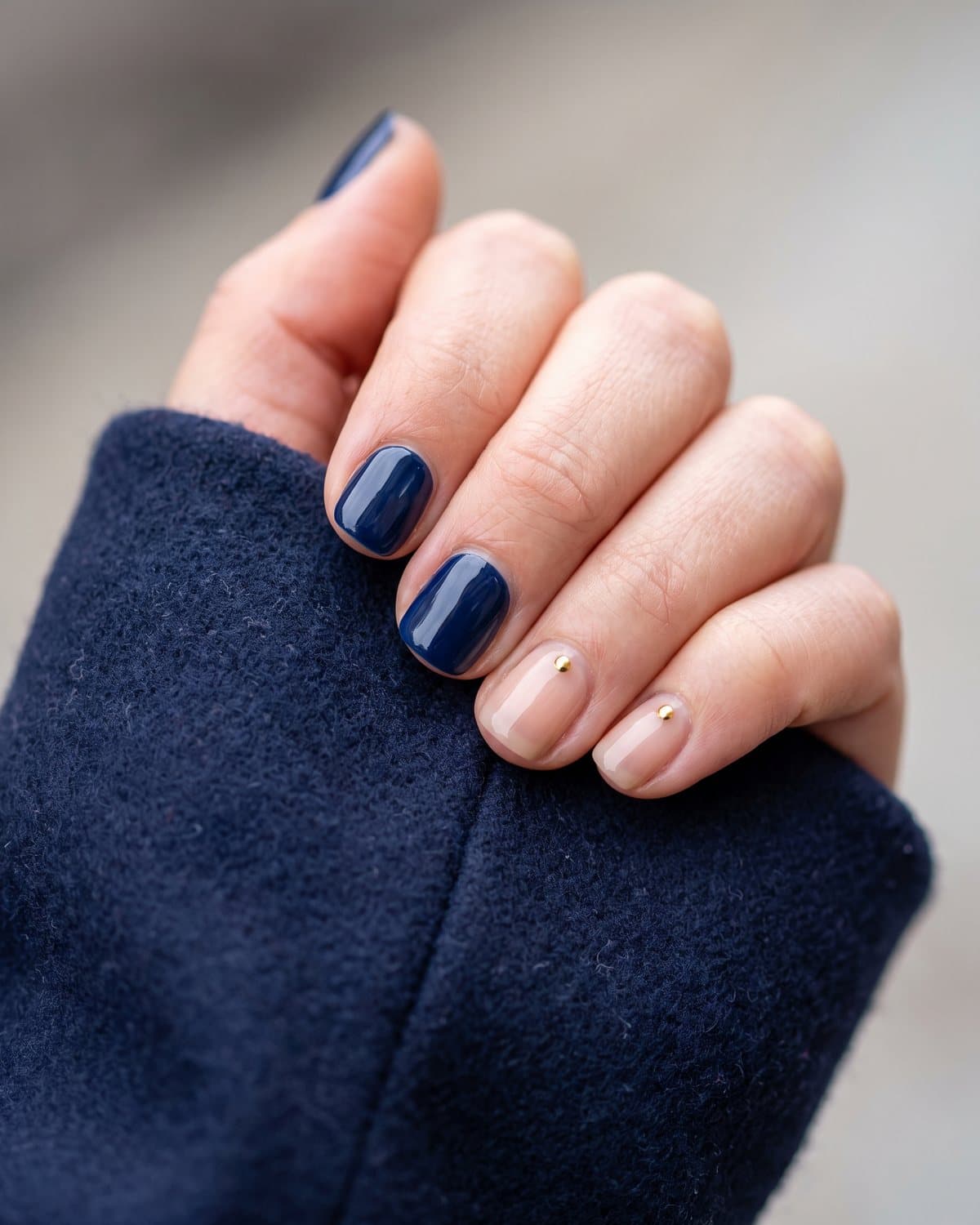 Short gel nails in glossy deep navy and sheer nude with a single tiny gold stud near the cuticle, resting on a navy wool coat