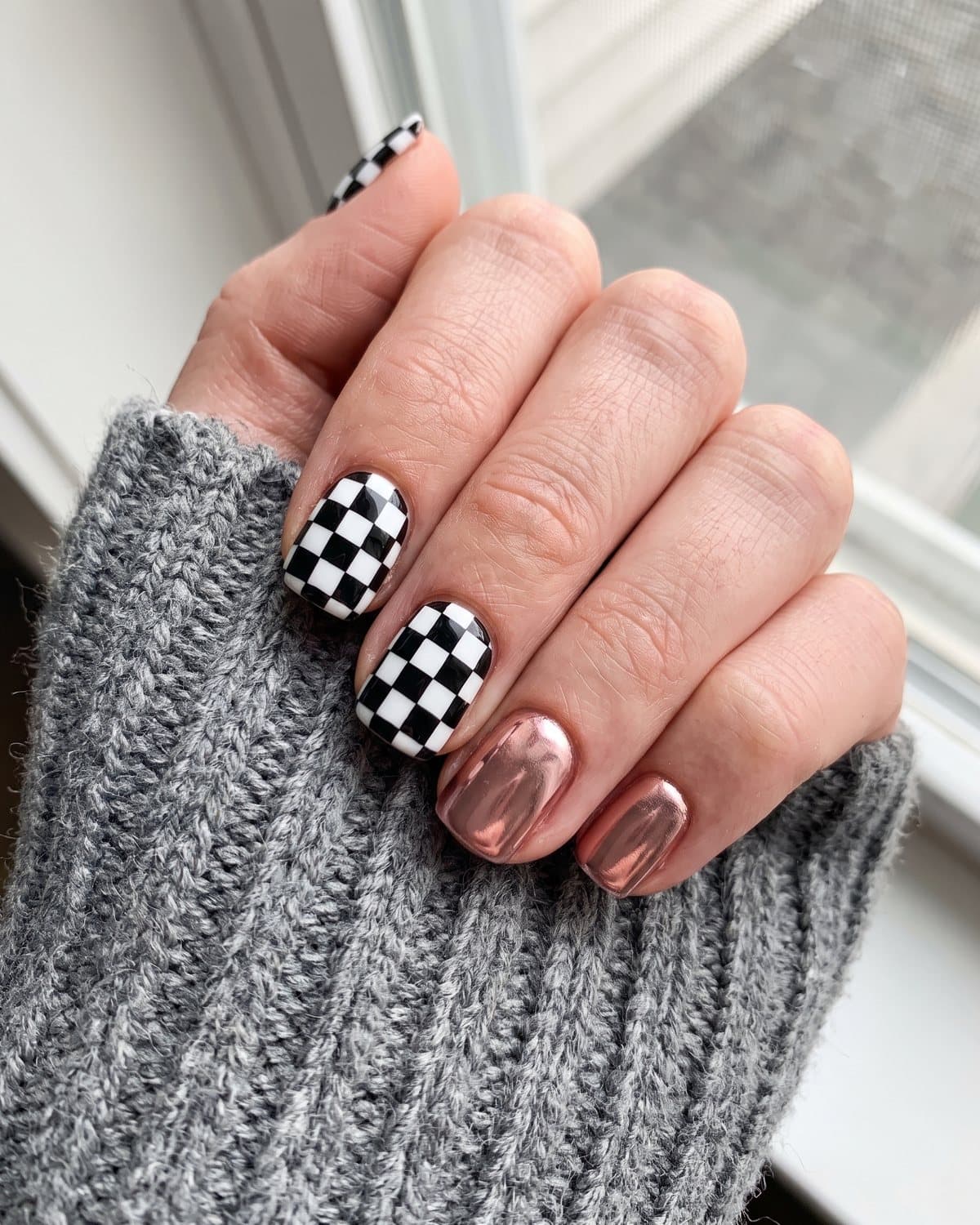 Short square nails with black and white checkerboard pattern and metallic rose gold chrome, on a grey knit