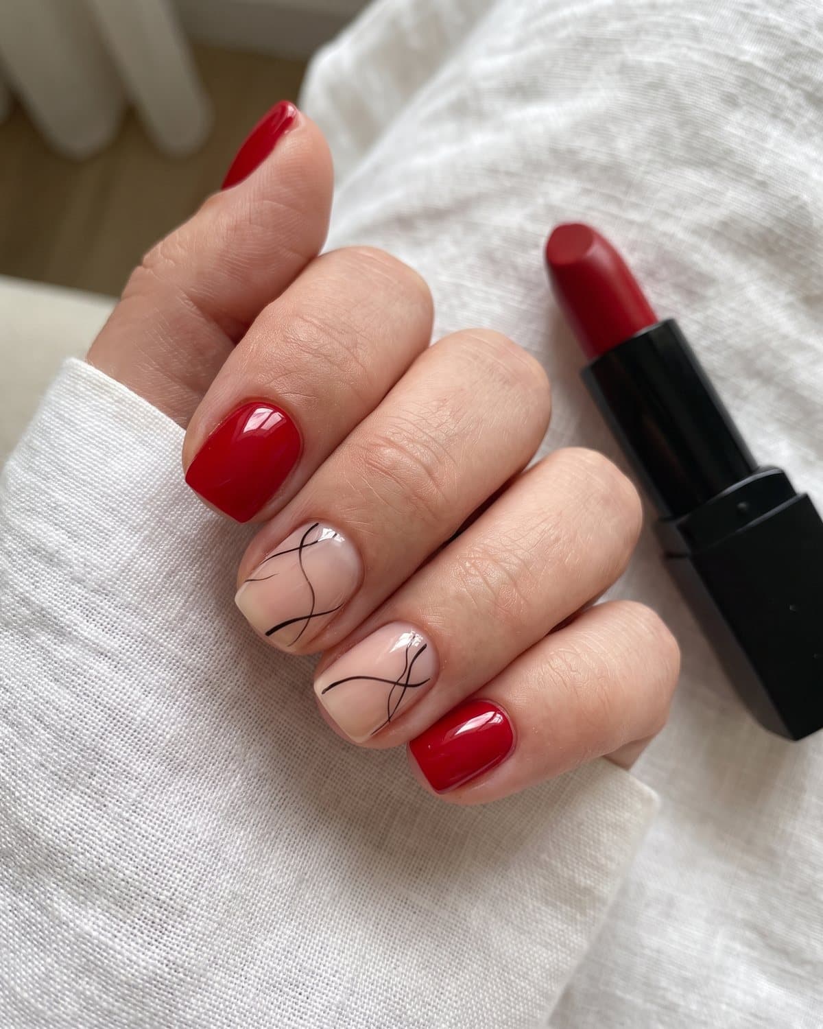 Short square nails in glossy classic red and sheer nude with thin minimalist black abstract lines, next to a red lipstick
