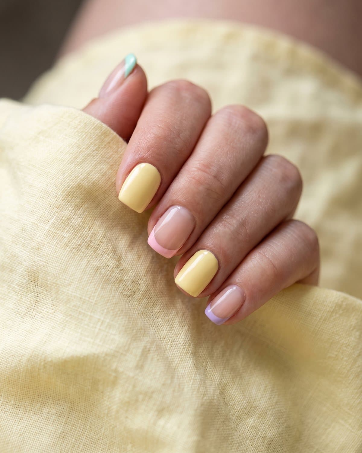 Short square nails with pastel French tips in mint, pink and lilac and soft butter yellow cream nails, on pale yellow linen