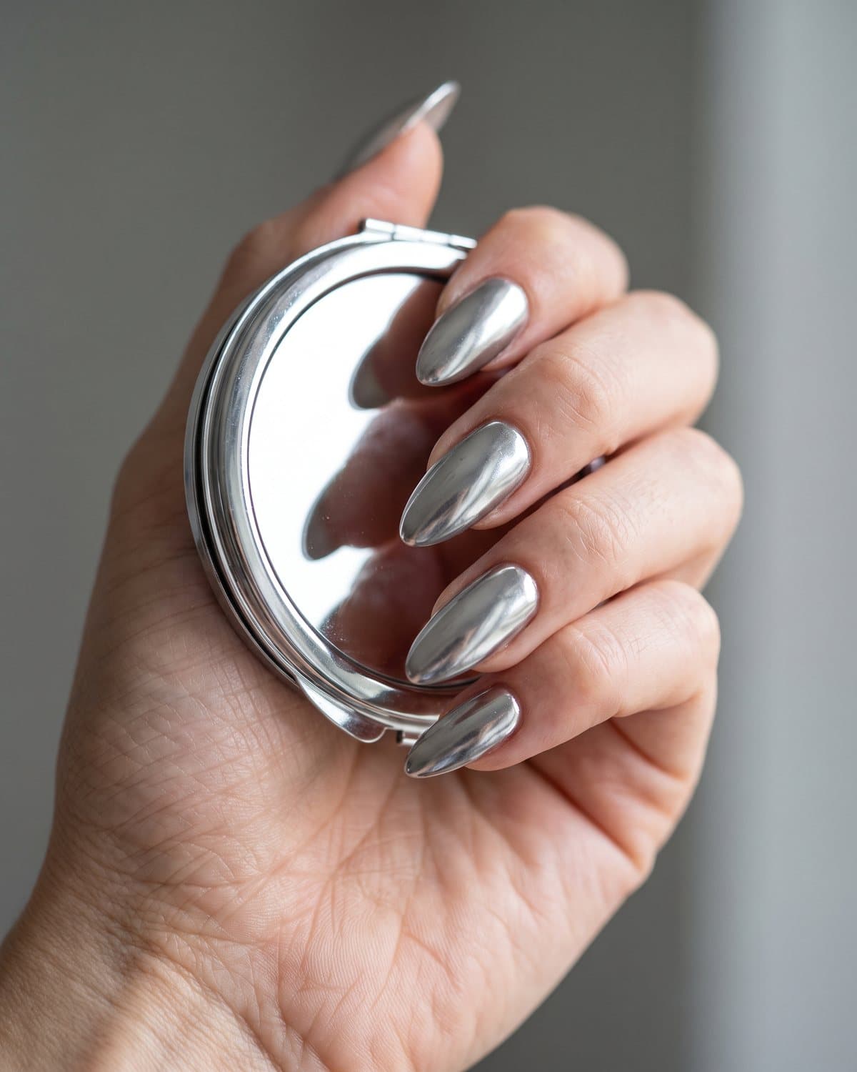 Long almond nails in highly reflective mirror silver chrome, holding a silver compact mirror against a grey background