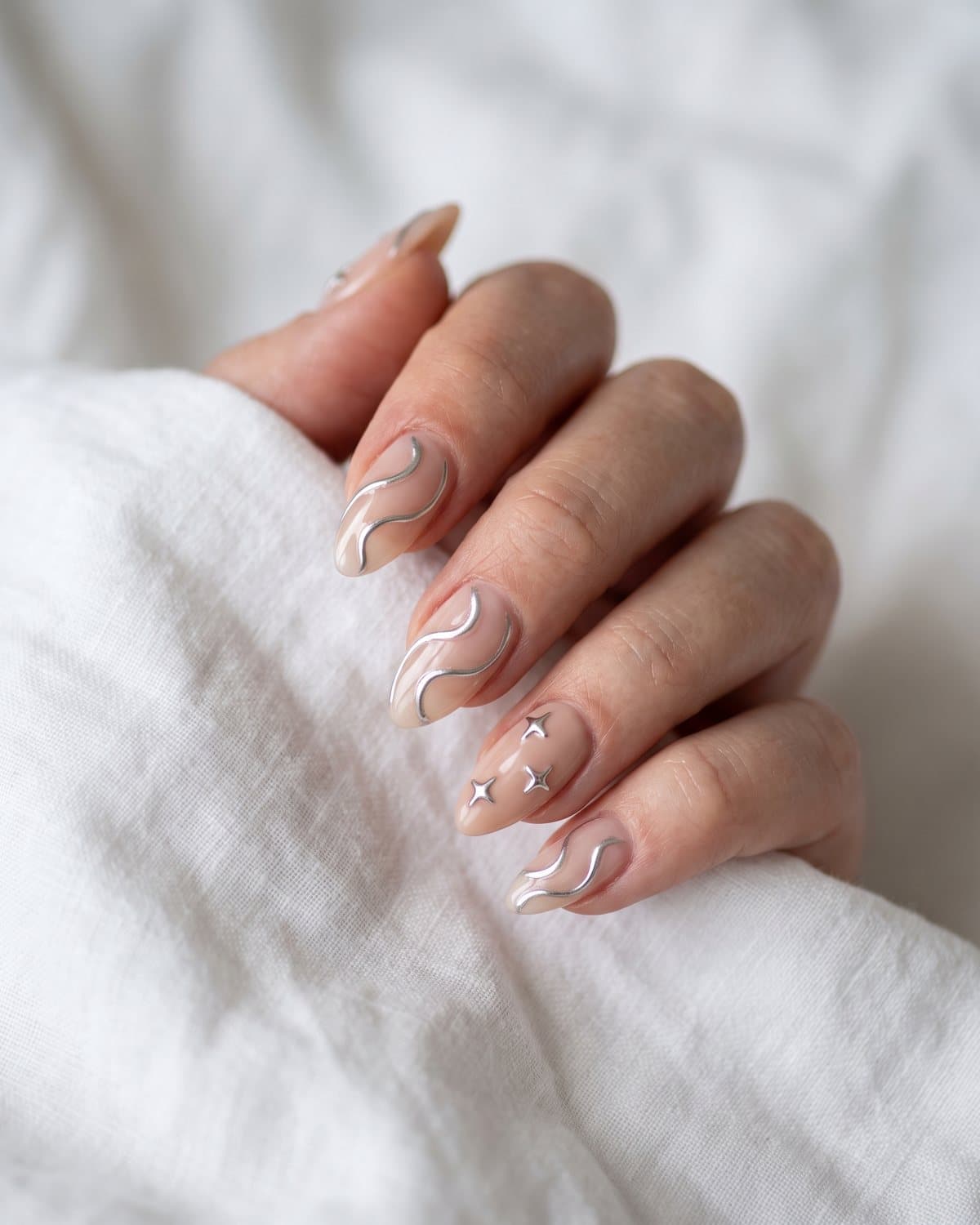 Almond nails with flowing silver chrome swirl lines on sheer nude and tiny raised silver chrome stars, on white linen