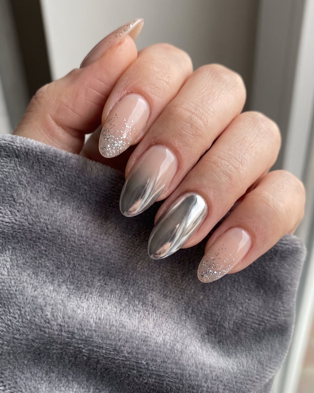 Almond nails with a silver chrome ombré from nude to mirror silver and nude nails with fine silver glitter fading from the tip, on grey velvet