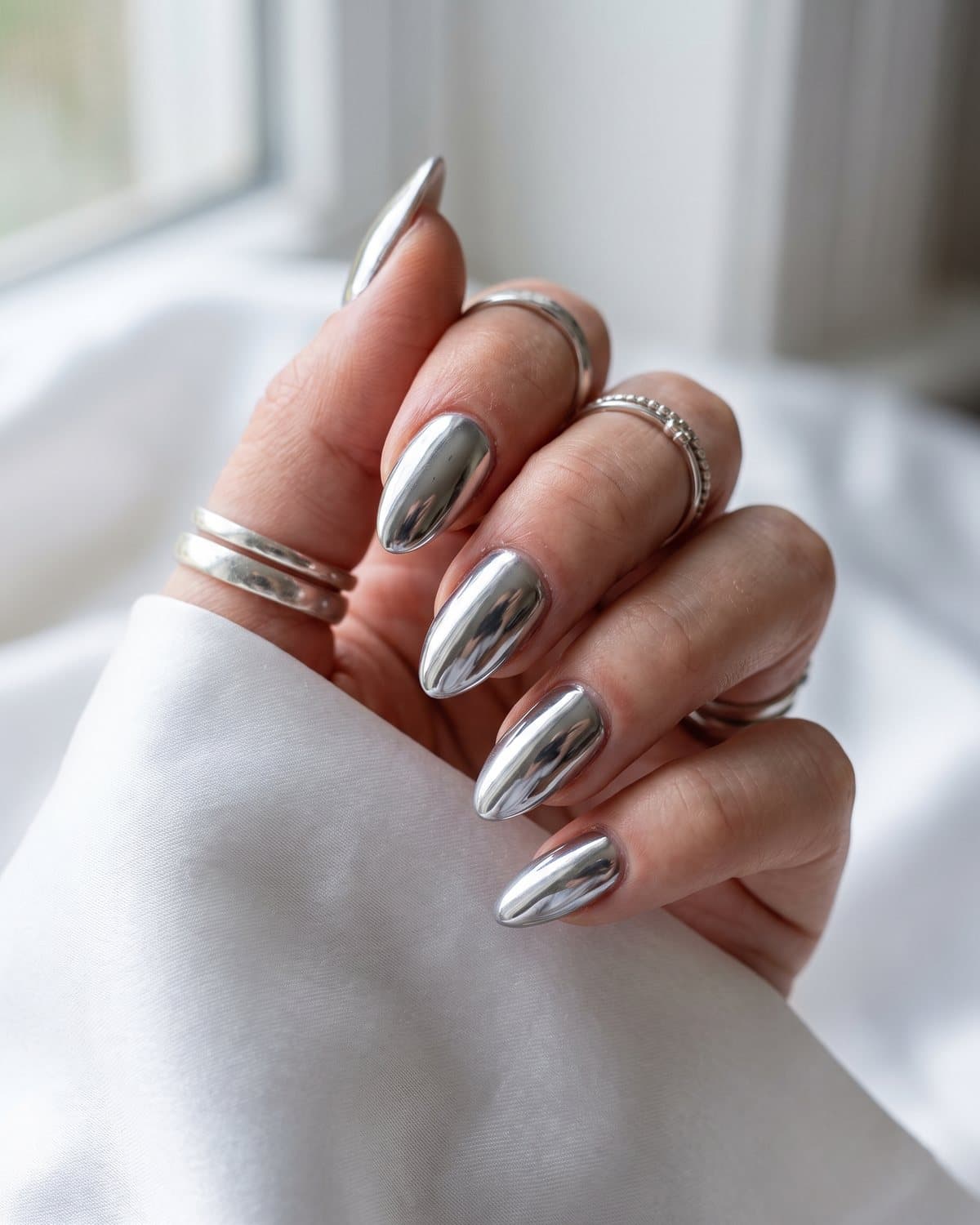 Almond nails in full mirror silver chrome with liquid metal reflections, resting on white satin with silver rings