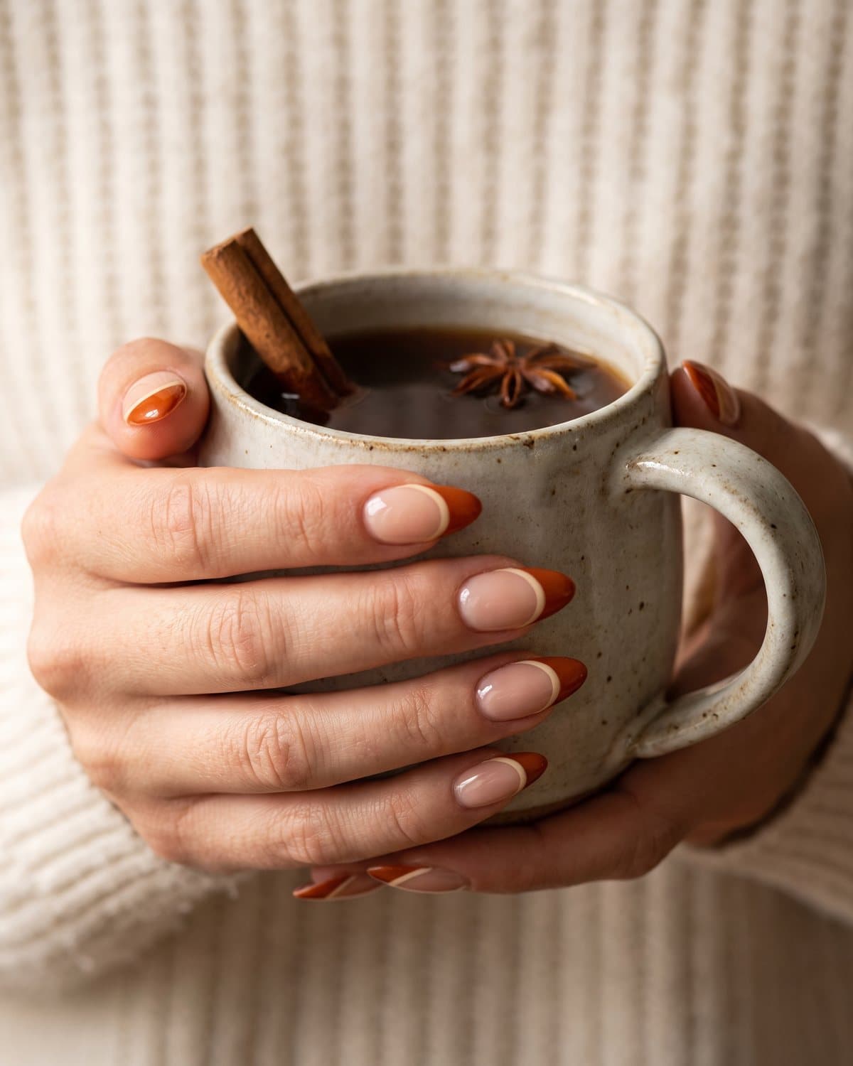 Almond nails with a sheer nude base and burnt orange French tips with a thin cream line, holding a ceramic mug of cider