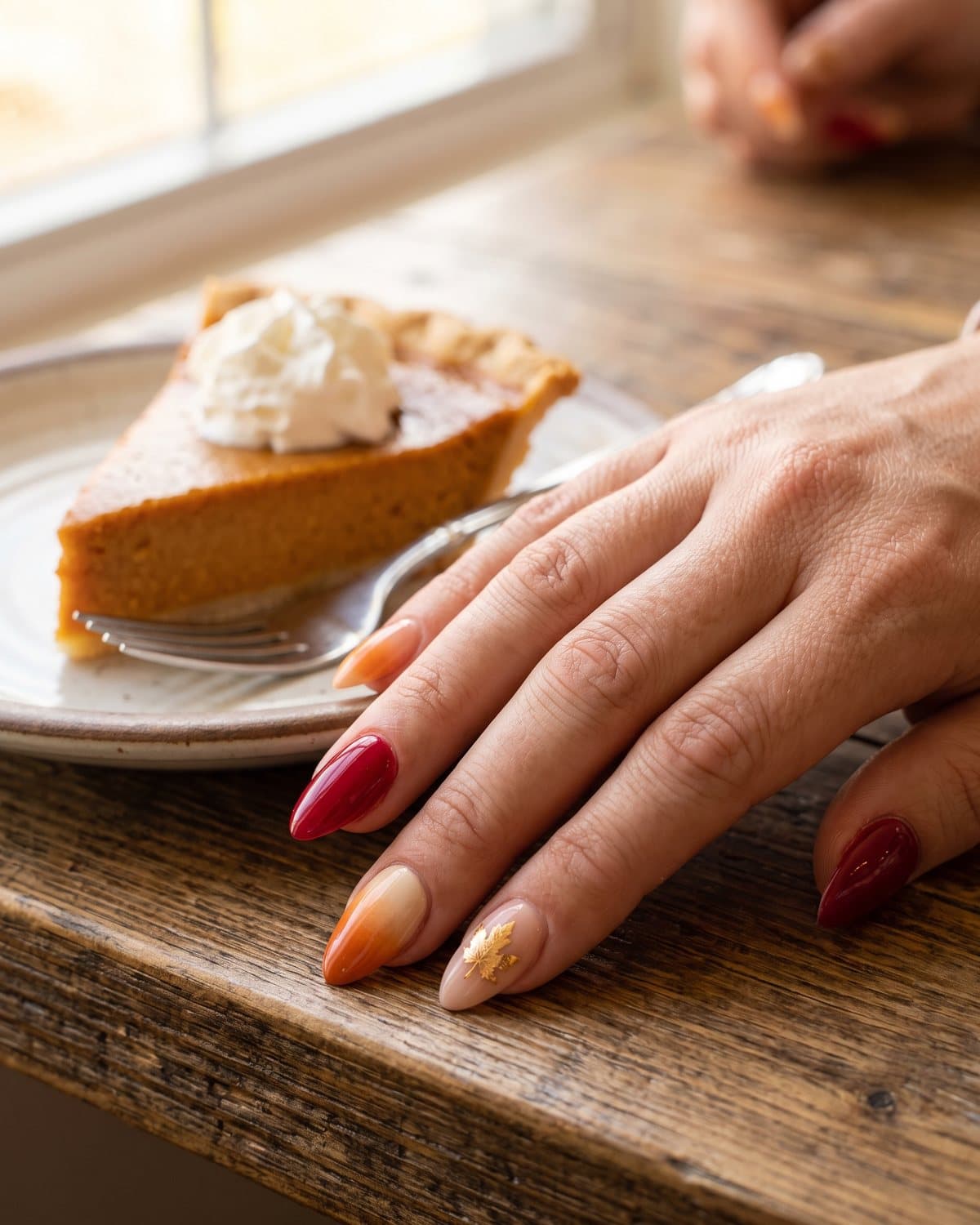 Almond nails with a cream-to-orange pumpkin pie ombré, glossy cranberry red and a gold leaf maple leaf, beside a slice of pumpkin pie