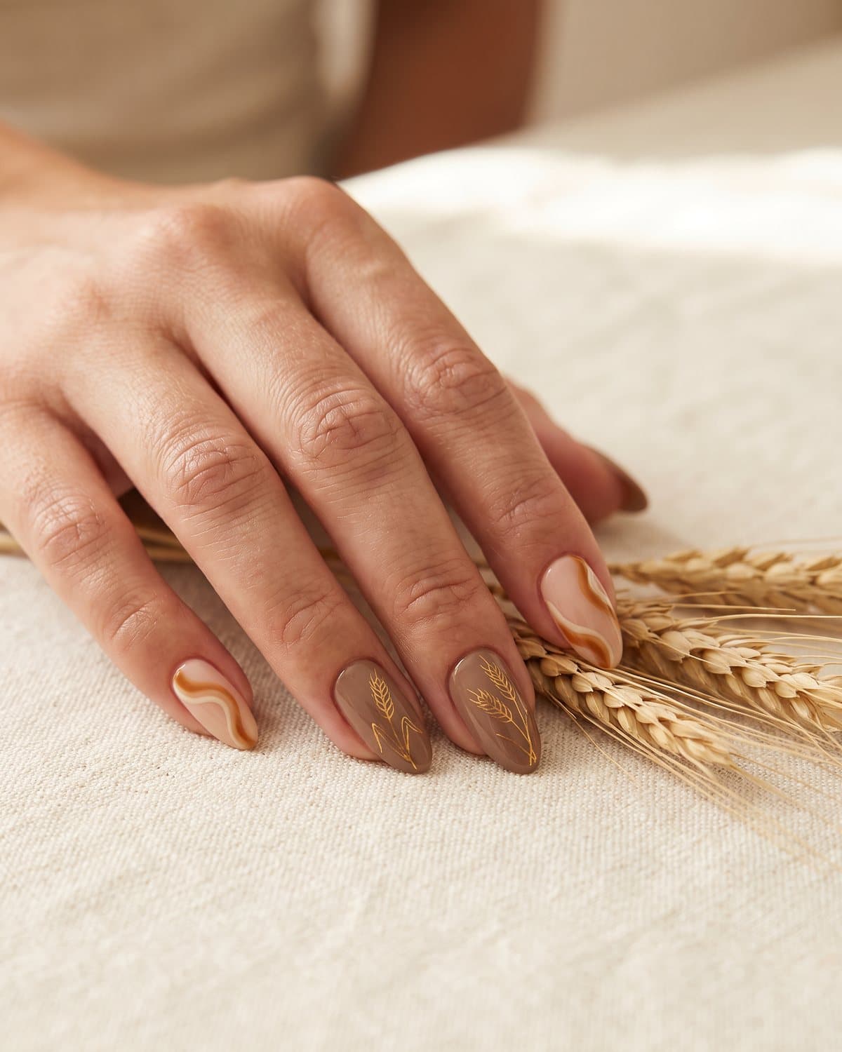 Almond nails with caramel and cream swirls and mocha nails with fine gold wheat stalk line art, on a cream tablecloth with dried wheat